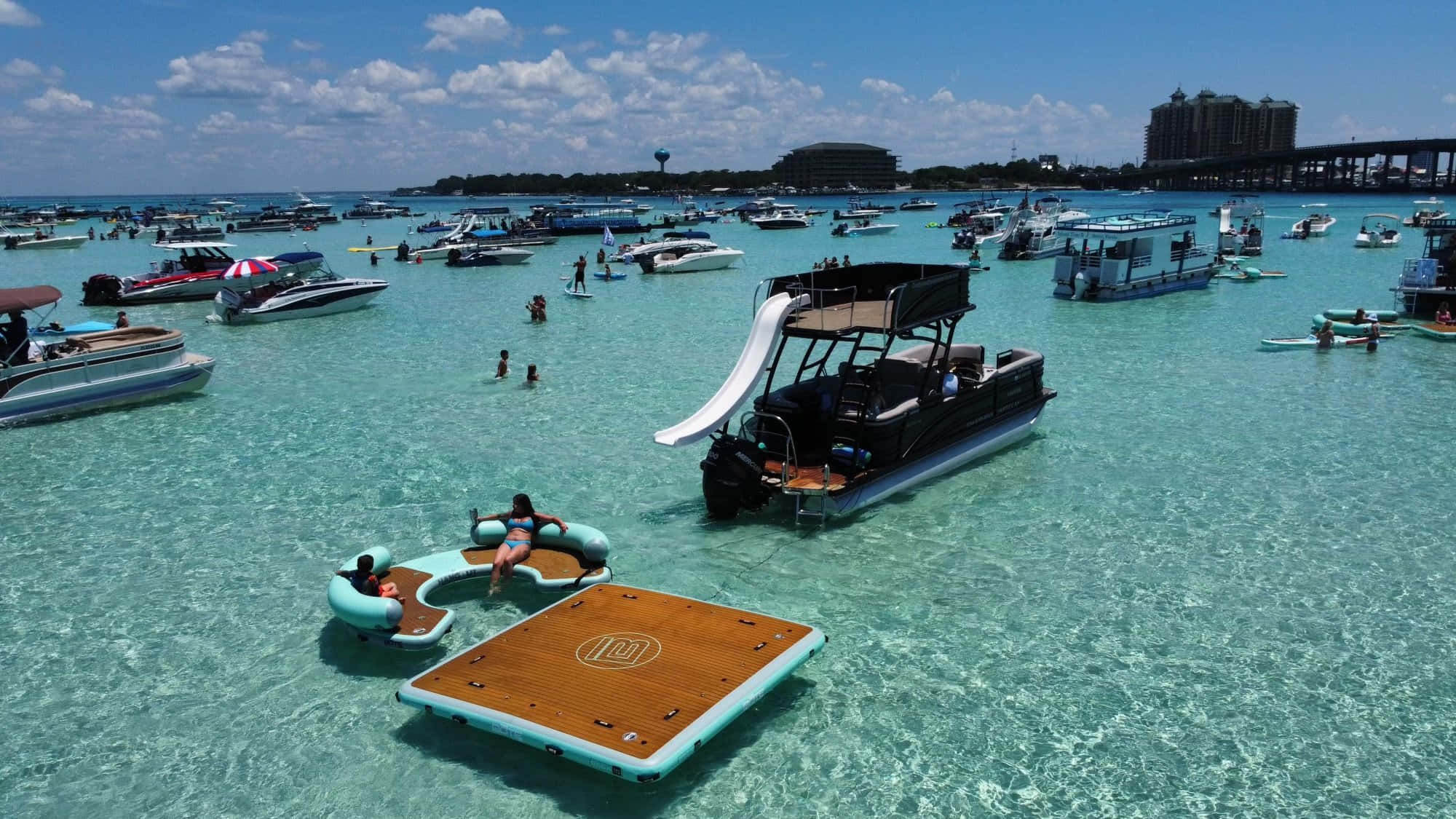 Crab Island 桌布 壁紙 — 從高空俯瞰，碧綠的海水中佈滿船隻和浮具，人們在遠處的度假村附近享受陽光普照的一天。
