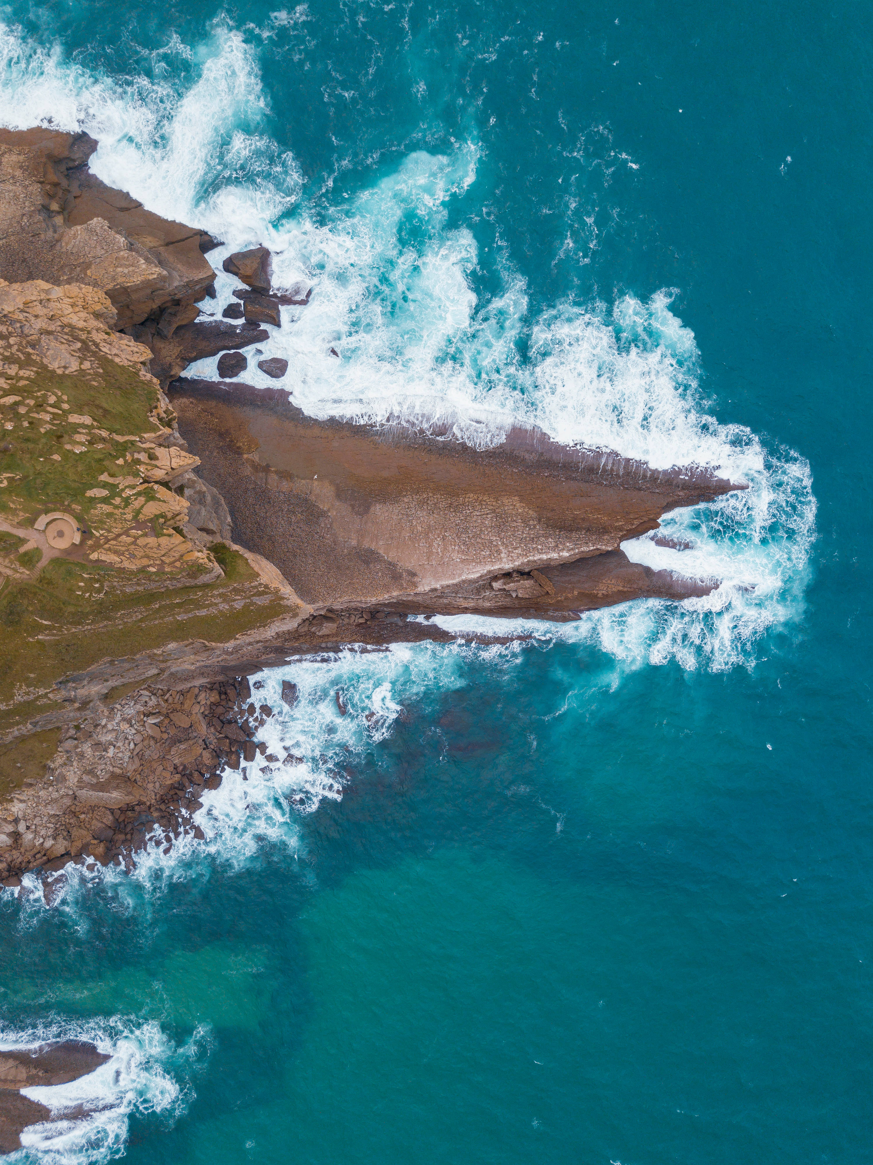 Cabo 桌布 壁紙 — 從高空俯瞰，碧綠色的海浪拍打著崎嶇的懸崖，懸崖上有綠色草叢和一個小型圓形結構。