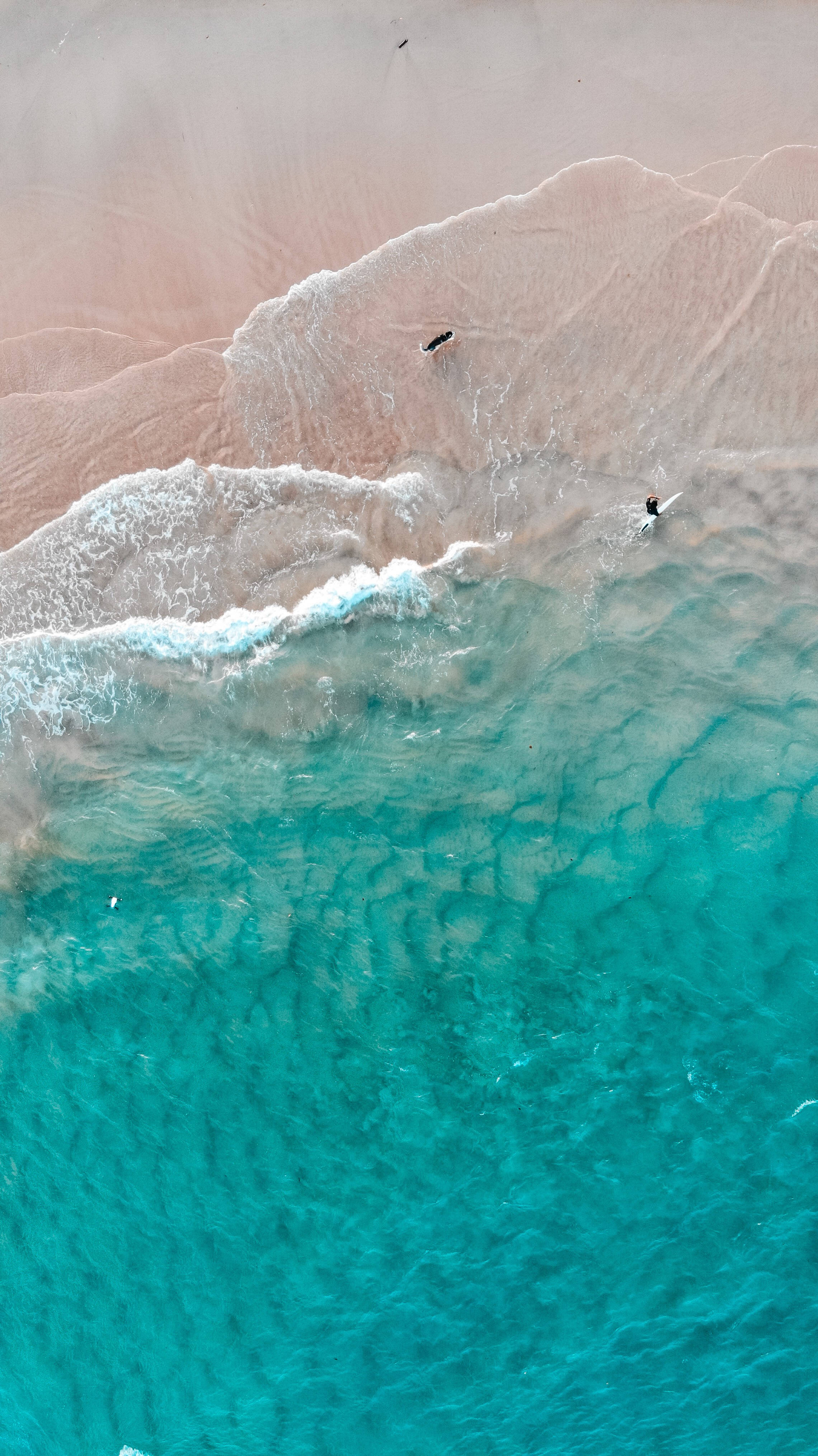 Aesthetic iPhone Xr 桌布 壁紙 — 從高空俯瞰，碧綠色的海浪輕輕拍打著淡粉色的沙灘，衝浪者在岸邊馳騁。