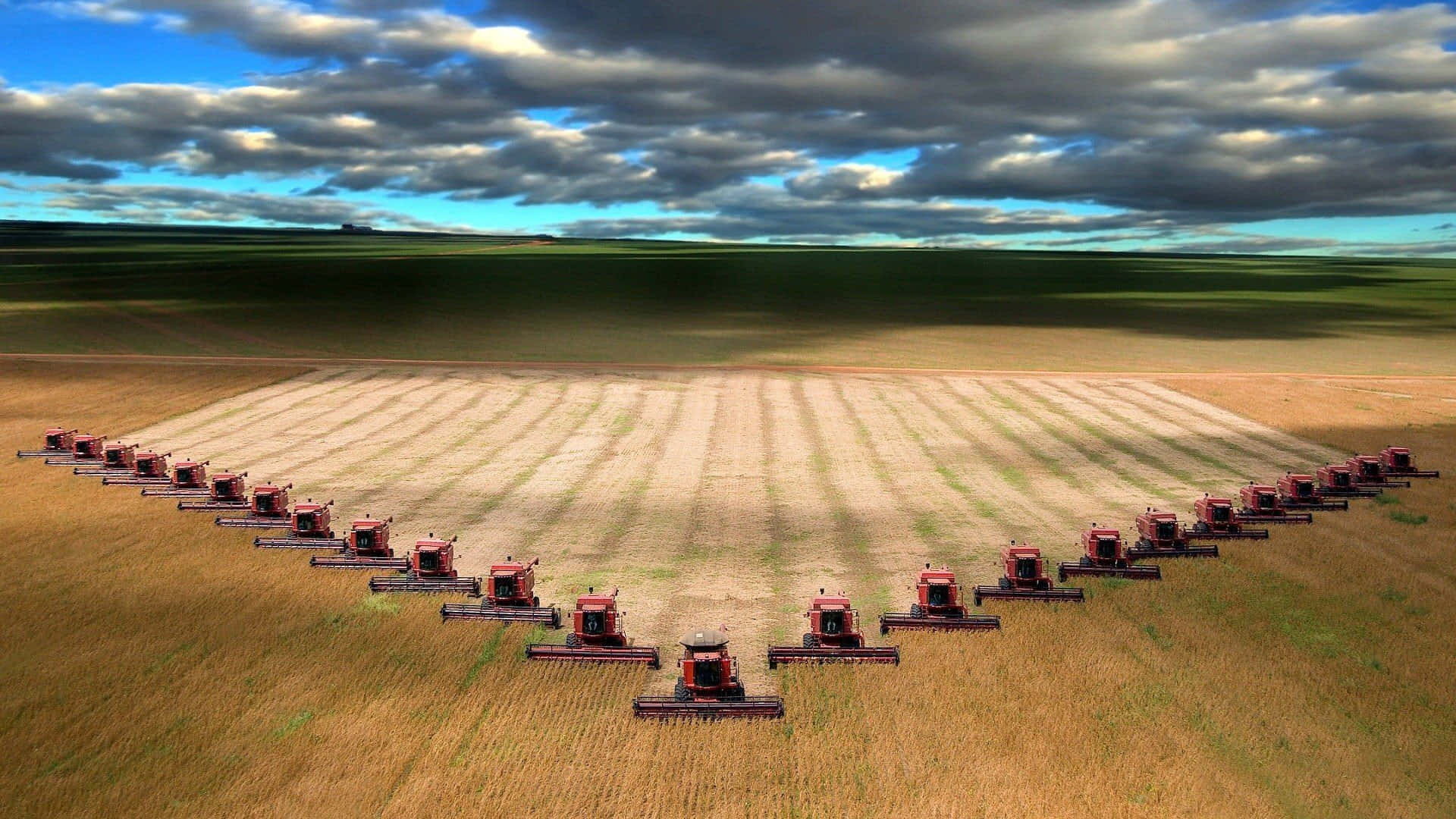 Agriculture 桌布 壁紙 — 從高空俯瞰，紅色收割機在金色麥田上形成一條曲線，背景是戲劇性的多雲天空。