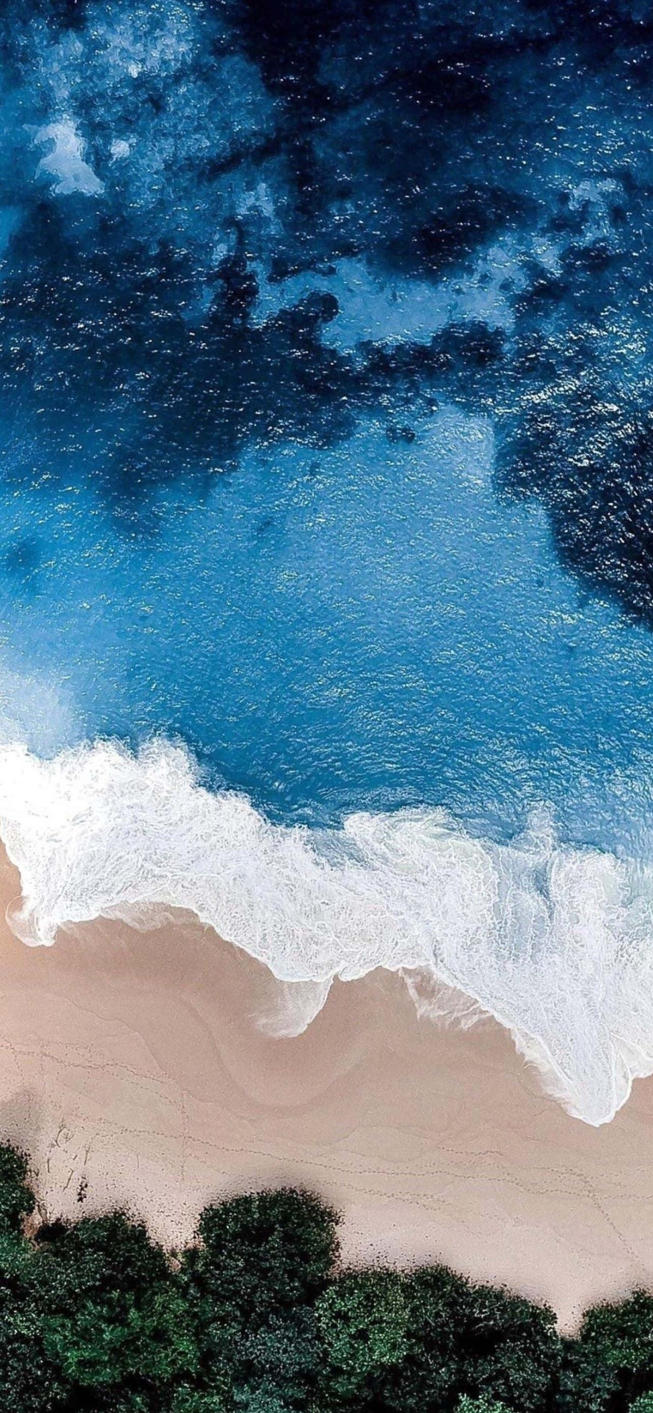 酷炫iPhone 桌布 壁紙 — 從高空俯瞰，綠松石色的海浪拍打著沙灘，沙灘邊緣長滿了茂密的綠色植物。