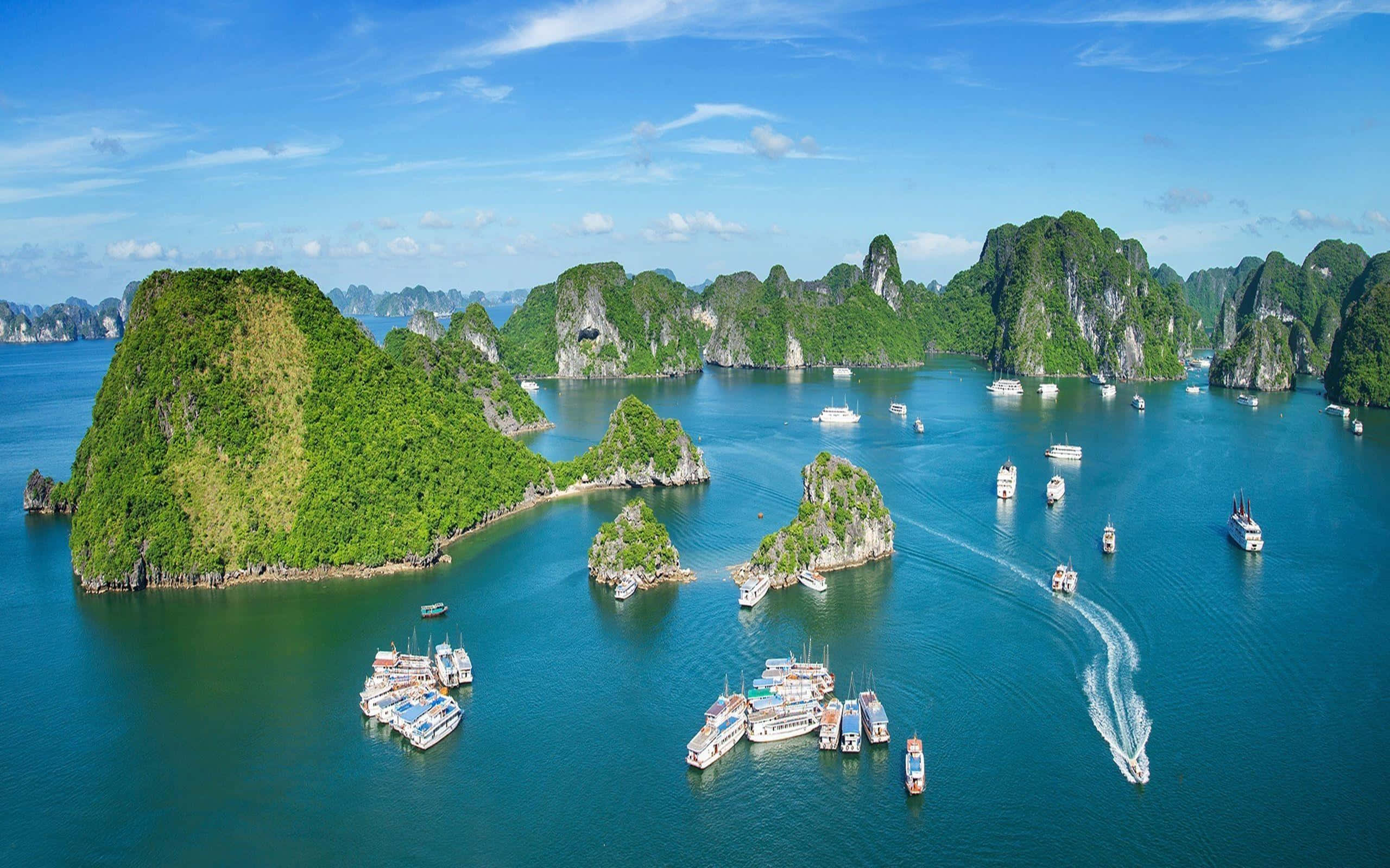 越南 桌布 壁紙 — 從高空俯瞰，翠綠的喀斯特島嶼從碧綠色的海水中升起，海面上點綴著觀光船，天空湛藍明亮。