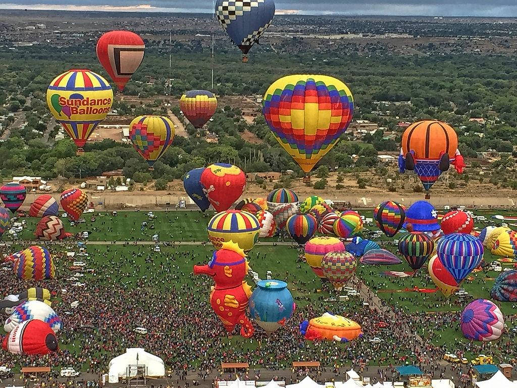 Balloon 桌布 壁紙 — 從高空俯瞰，色彩繽紛的熱氣球在擠滿人群的節日場地上方翱翔，周圍有綠樹和遠處的城鎮。