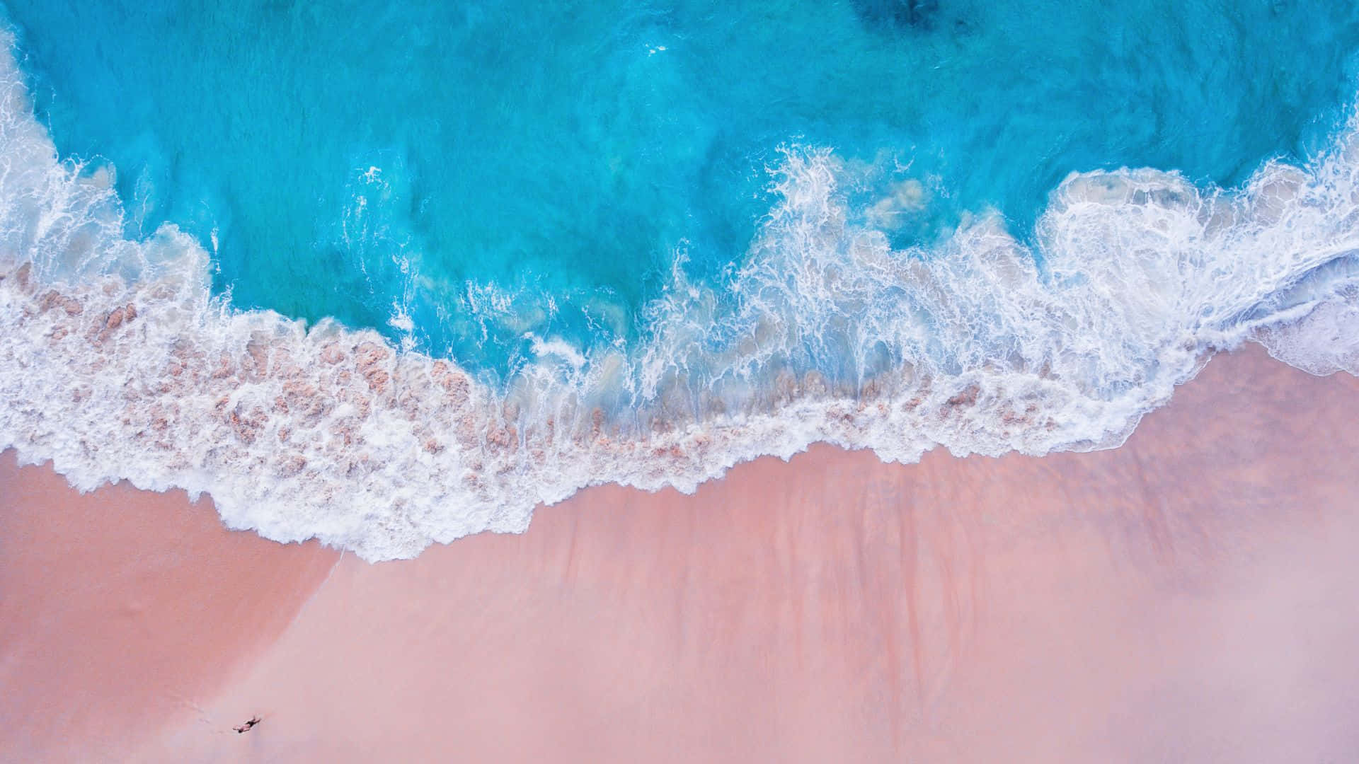 Pink Beach 桌布 壁紙 — 從高空俯瞰，藍綠色海浪拍打柔軟粉紅沙灘，泛起白色泡沫。