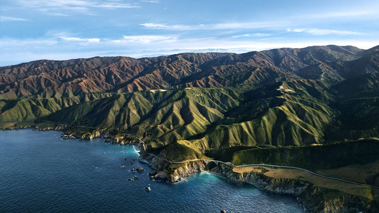 山脈 桌布 壁紙 — 從高空俯瞰，蜿蜒的沿海高速公路緊貼著崎嶇的綠色山脈，旁邊是深藍色的海洋，天空晴朗。