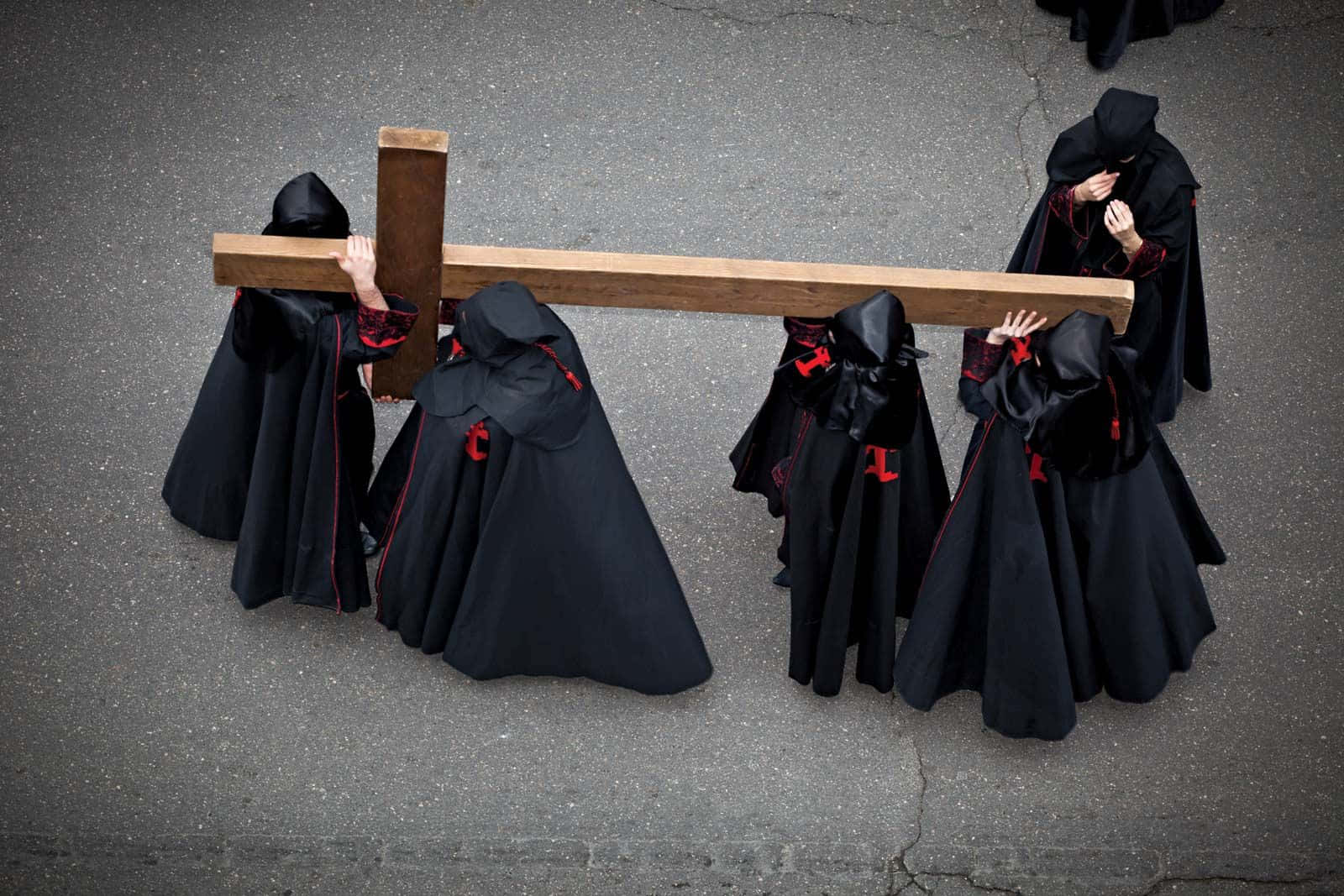Good Friday 桌布 壁紙 — 從高空俯瞰，身穿黑色紅邊長袍、戴著頭罩的人物，正抬著一個大木十字架走過龜裂的瀝青路面。