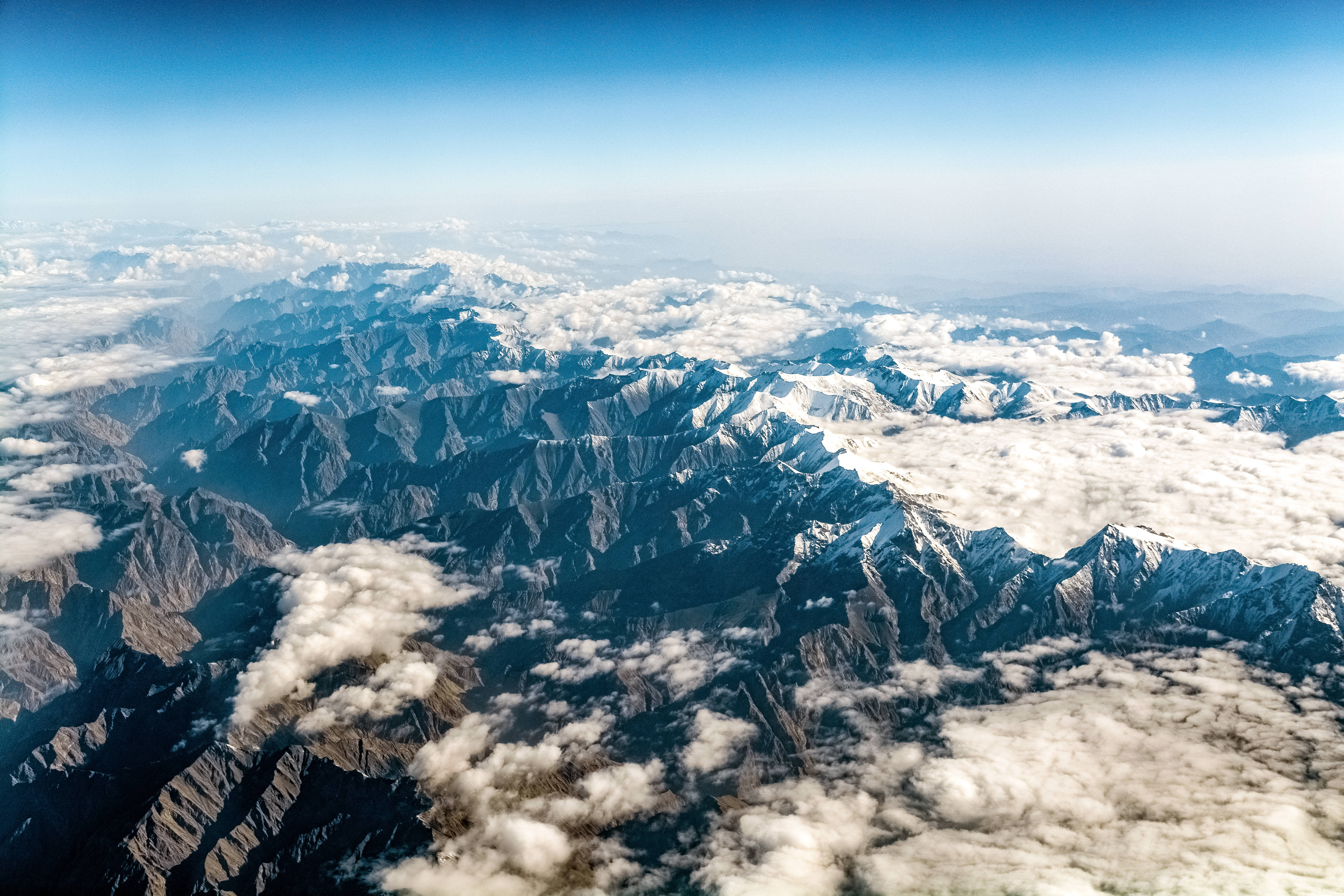 地形 桌布 壁紙 — 從高空俯瞰，鋸齒狀的積雪山峰在清澈藍天下穿過蓬鬆的白雲。
