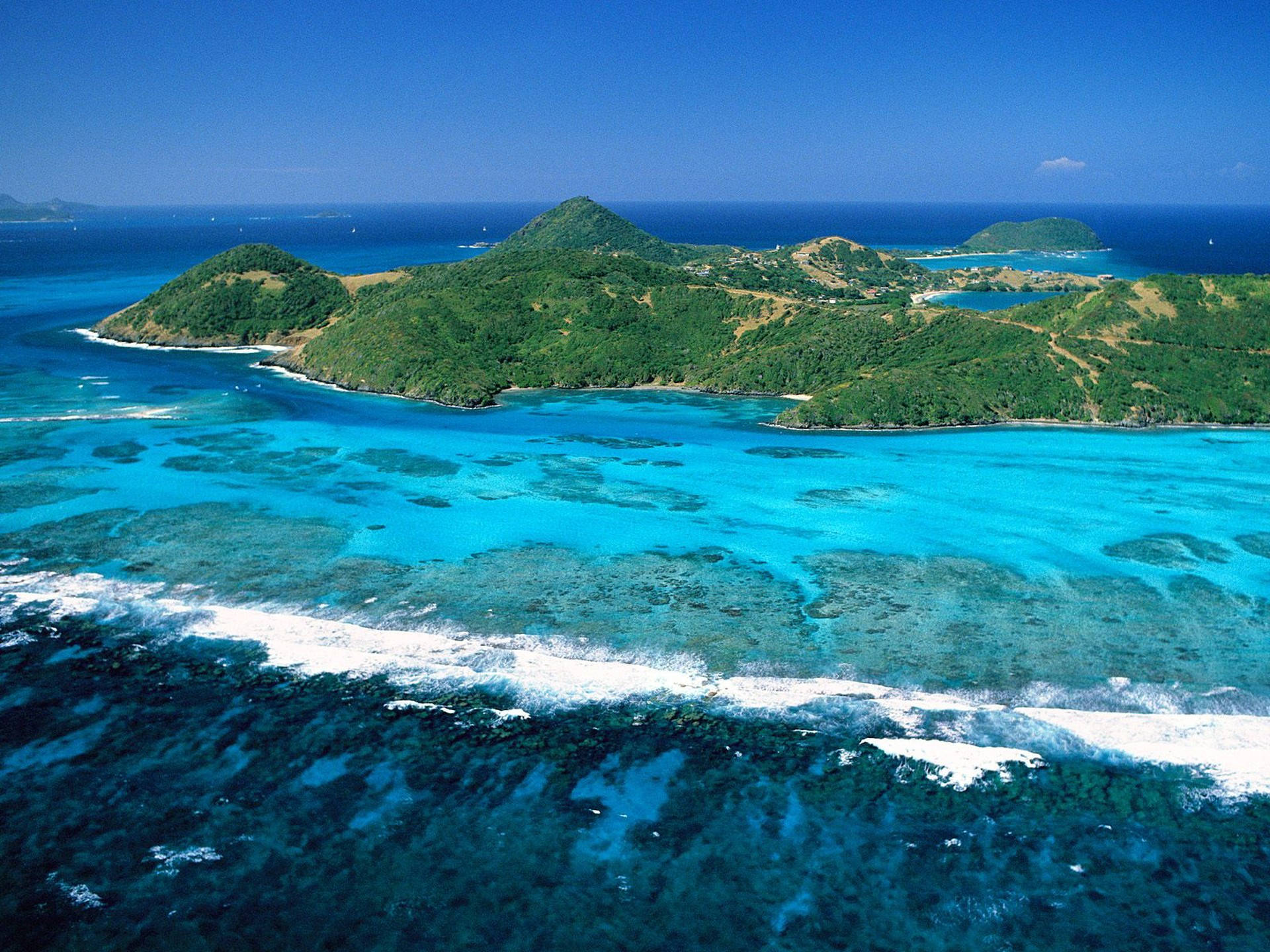 Caribbean 桌布 壁紙 — 從高空俯瞰，鬱鬱蔥蔥的綠色島嶼被充滿活力的碧綠海水和珊瑚礁環繞，上方是清澈的藍天。