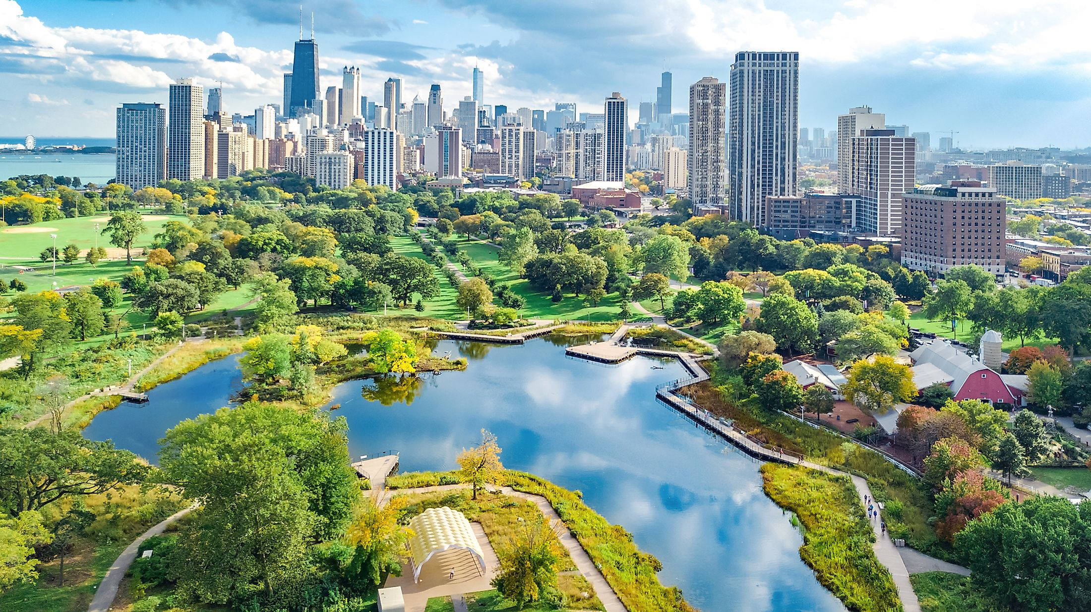 Beautiful Nature Photography 桌布 壁紙 — 從高空俯瞰一個充滿活力的城市公園，蜿蜒的湖泊被秋樹和現代摩天大樓環繞，天空部分多雲。