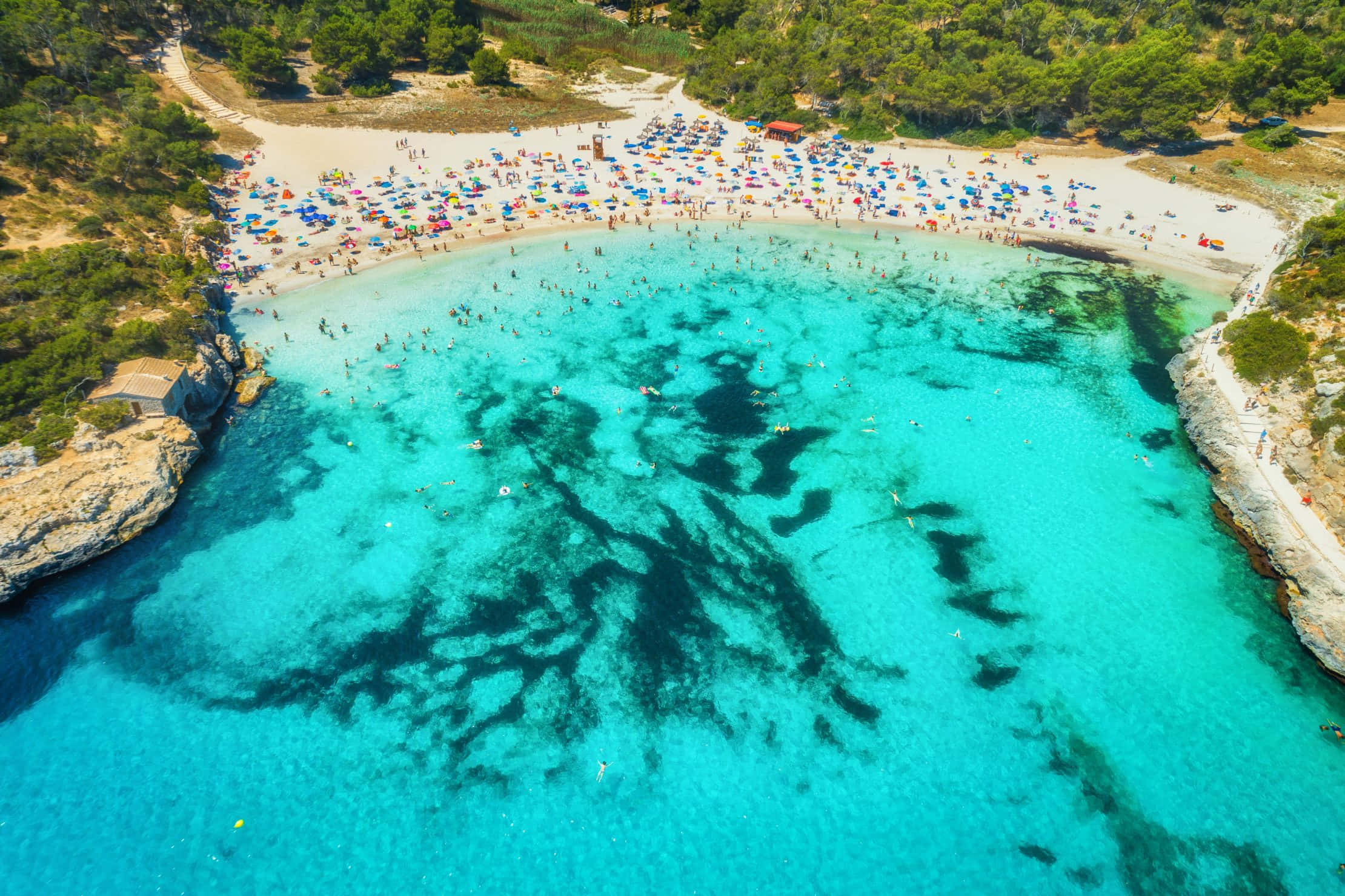 Spanish Beach 桌布 壁紙 — 從高空俯瞰一個充滿活力的綠松石色海灣，沙灘上有游泳者和日光浴者，四周被綠色懸崖環繞。