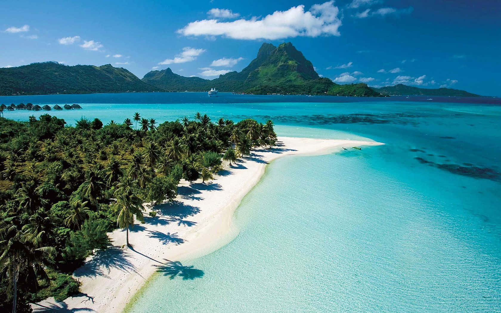 Bahamas Beach 桌布 壁紙 — 從高空俯瞰一個原始熱帶島嶼，擁有白色沙灘、棕櫚樹和碧綠潟湖，四周被茂密的綠色山脈環繞，天空湛藍明亮。
