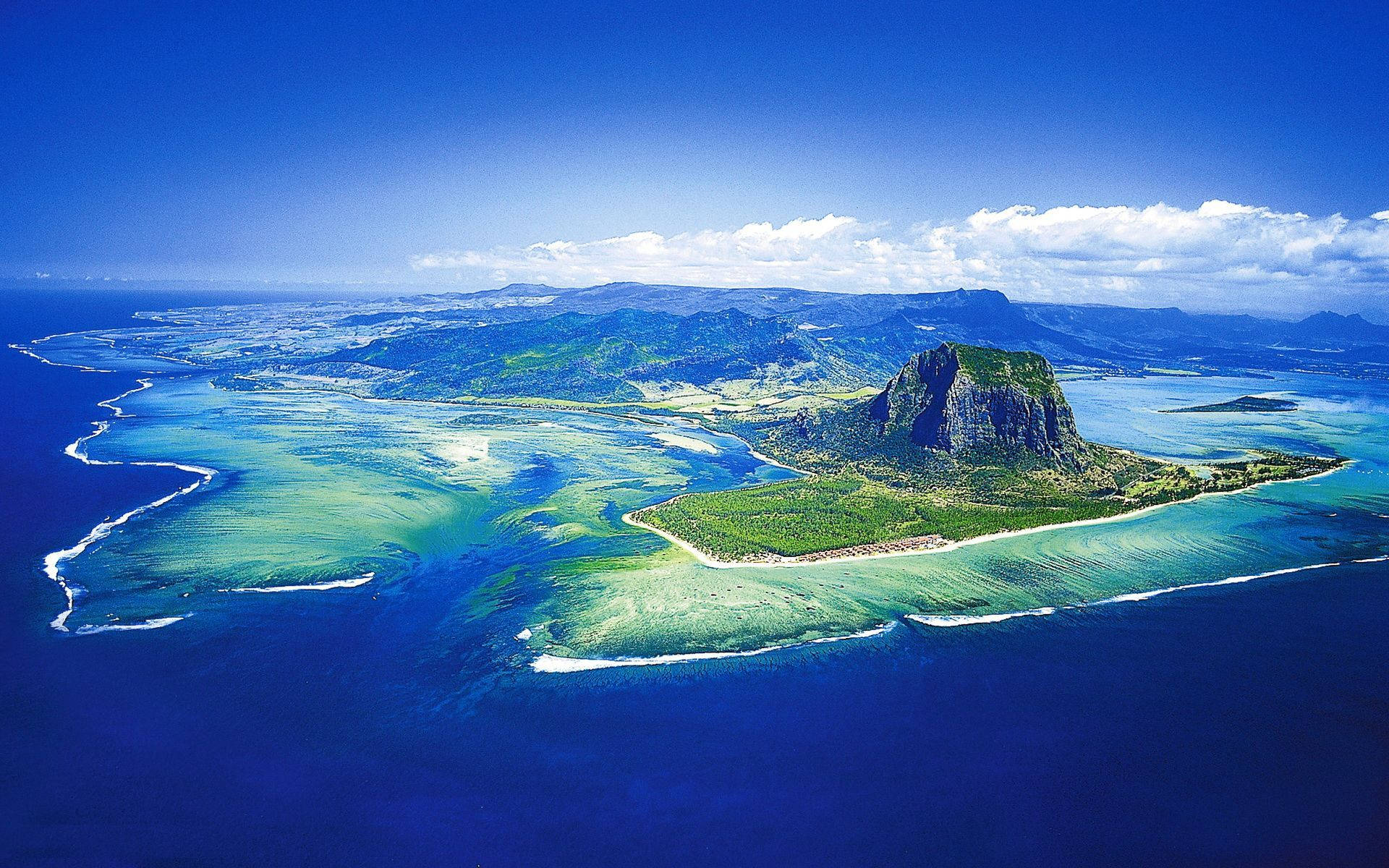 Mauritius 桌布 壁紙 — 從高空俯瞰一個鬱鬱蔥蔥的綠色島嶼，島上有一個突出的岩石山峰，周圍是充滿活力的碧綠海水和珊瑚礁，天空清澈湛藍。