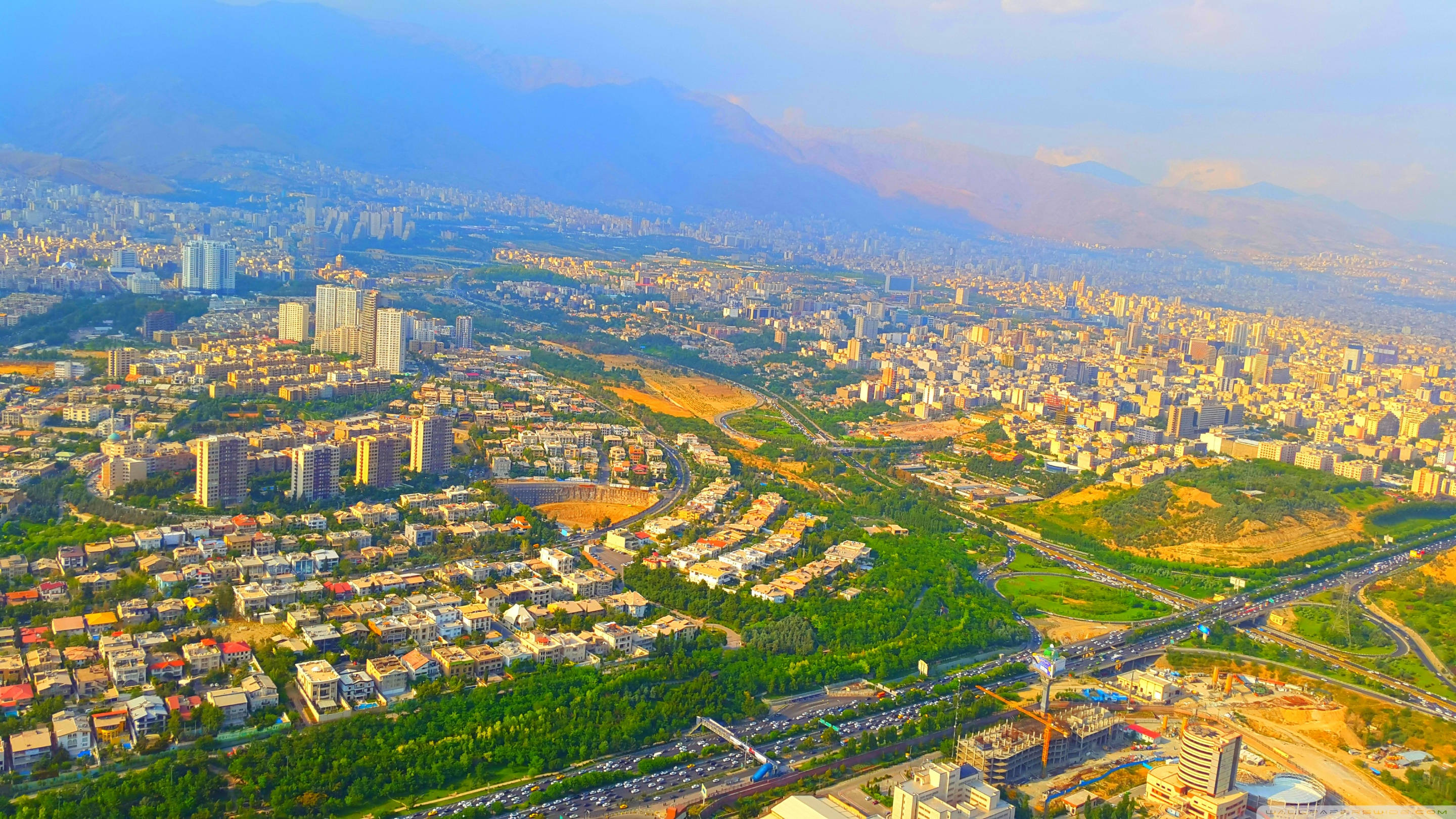 Tehran 桌布 壁紙 — 從高空俯瞰一座坐落在山谷中的廣闊城市，背景是沐浴在金色陽光下的山脈。
