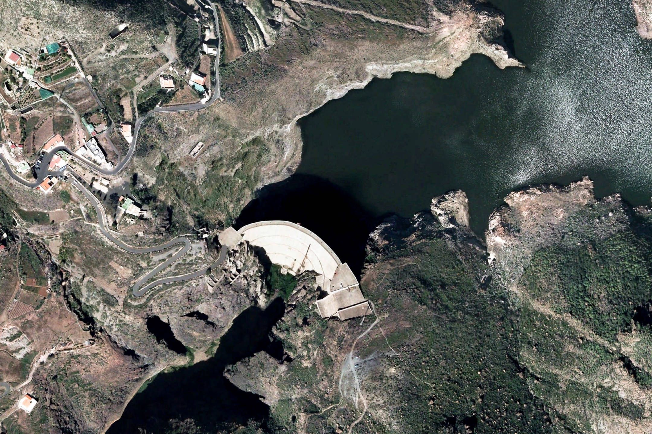 Google 地球 桌布 壁紙 — 從高空俯瞰一座巨大的弧形水壩，在崎嶇的山地地形中攔截著一個黑暗的水庫，周圍有蜿蜒的道路和散落的建築物。
