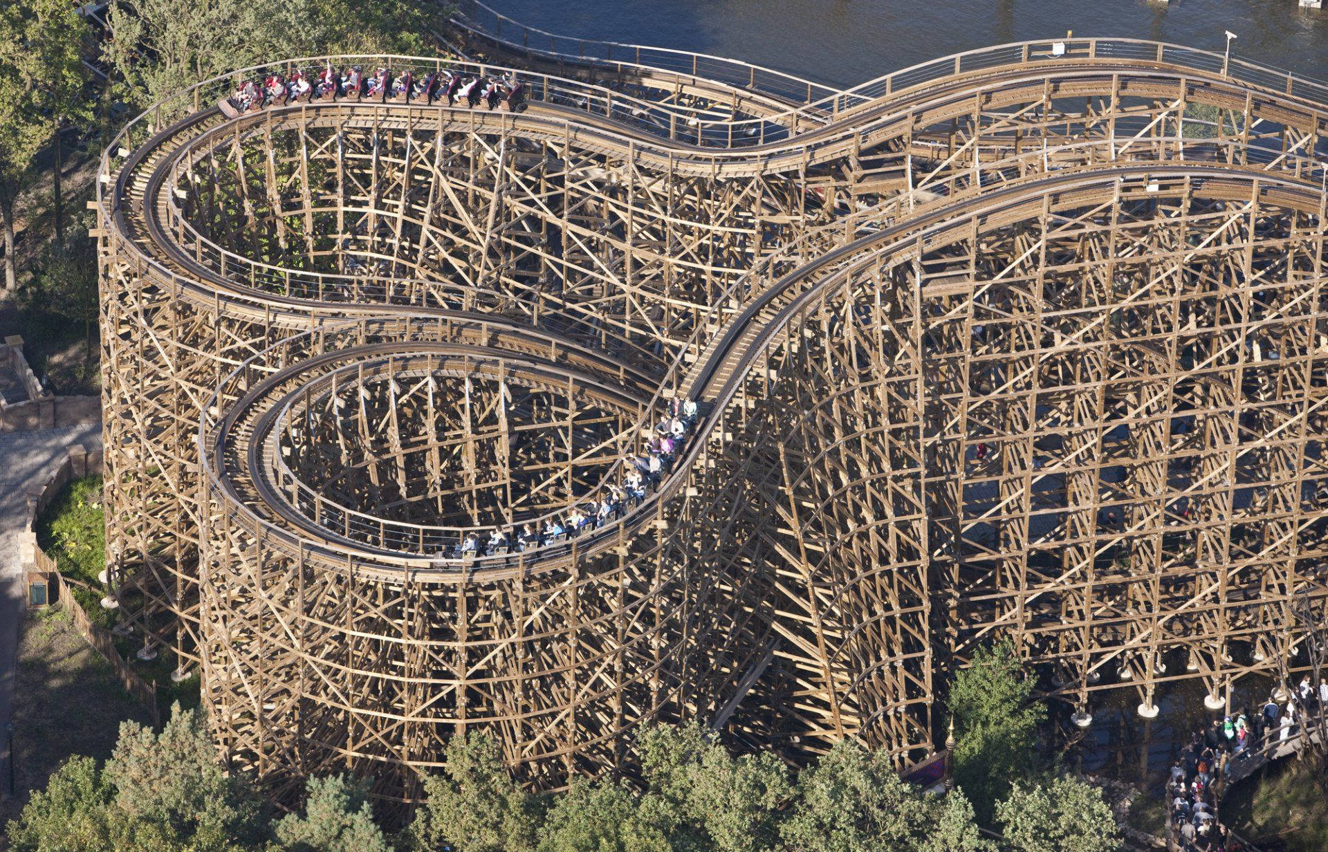 Roller Coaster 桌布 壁紙 — 從高空俯瞰一座巨大的木製雲霄飛車，蜿蜒穿過河邊茂密的綠樹，車上有乘客。