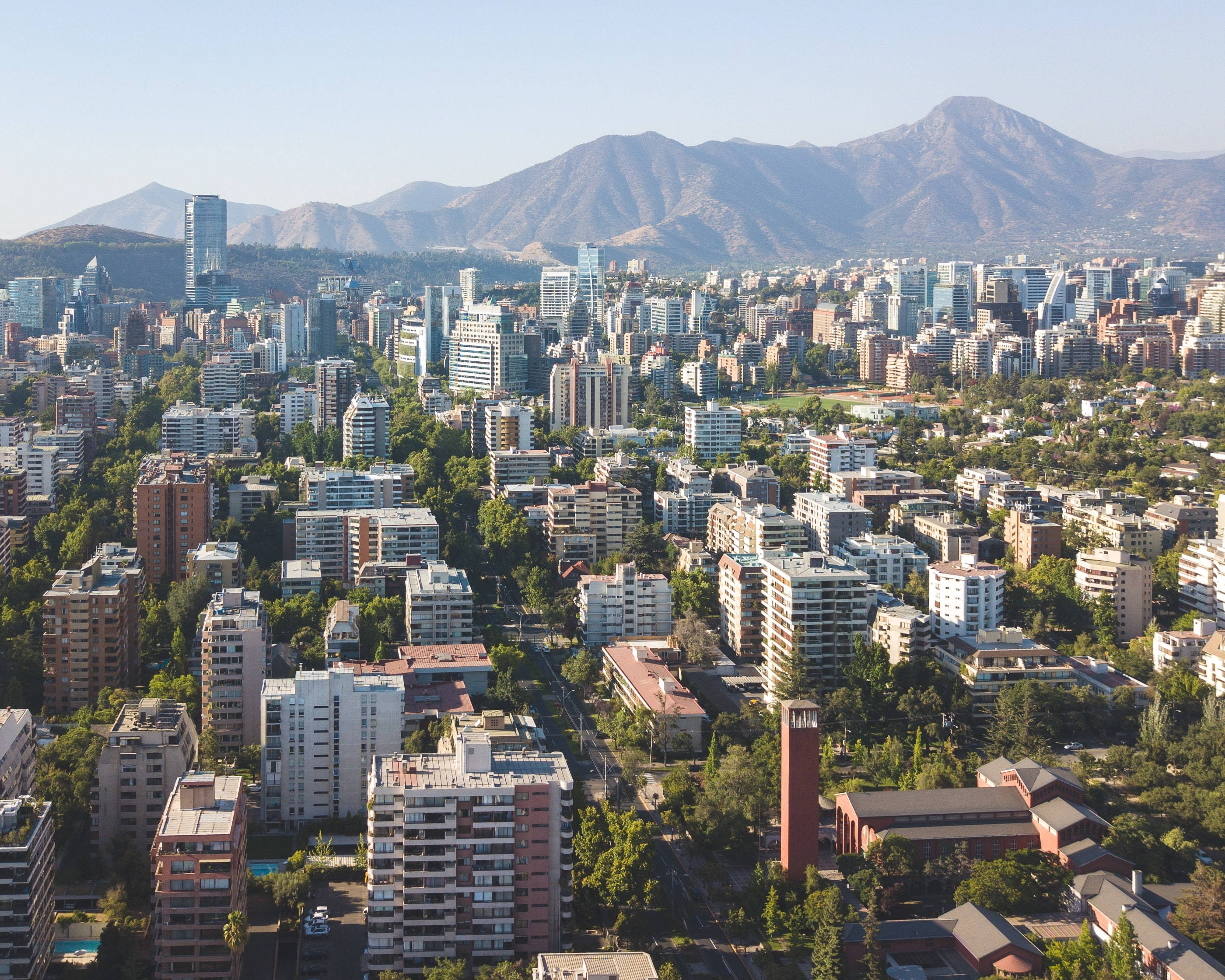 山景 桌布 壁紙 — 從高空俯瞰一座廣闊的城市，現代建築與茂密綠植交織，背景是雄偉的山脈，天空湛藍清澈。