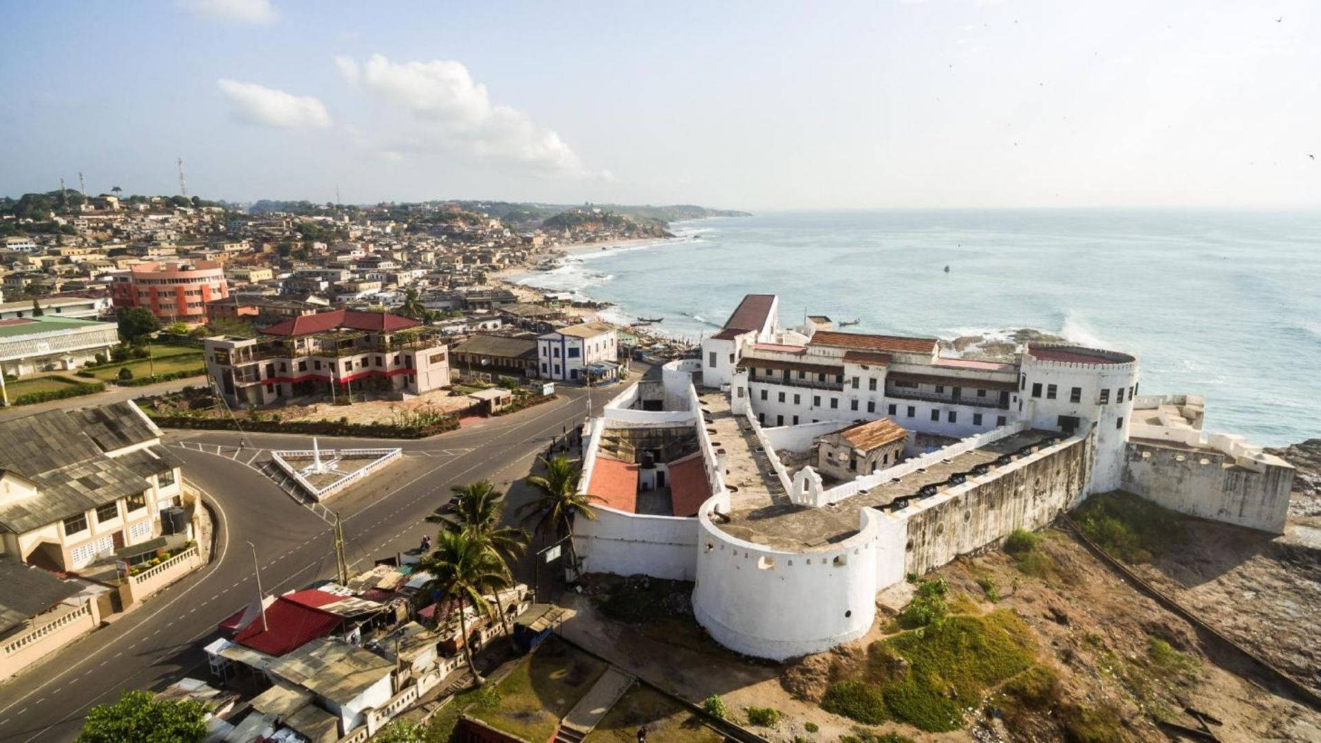 Ghana 桌布 壁紙 — 從高空俯瞰一座歷史悠久的白色堡壘，俯瞰著沿海城市和碧綠的海洋，天空晴朗。
