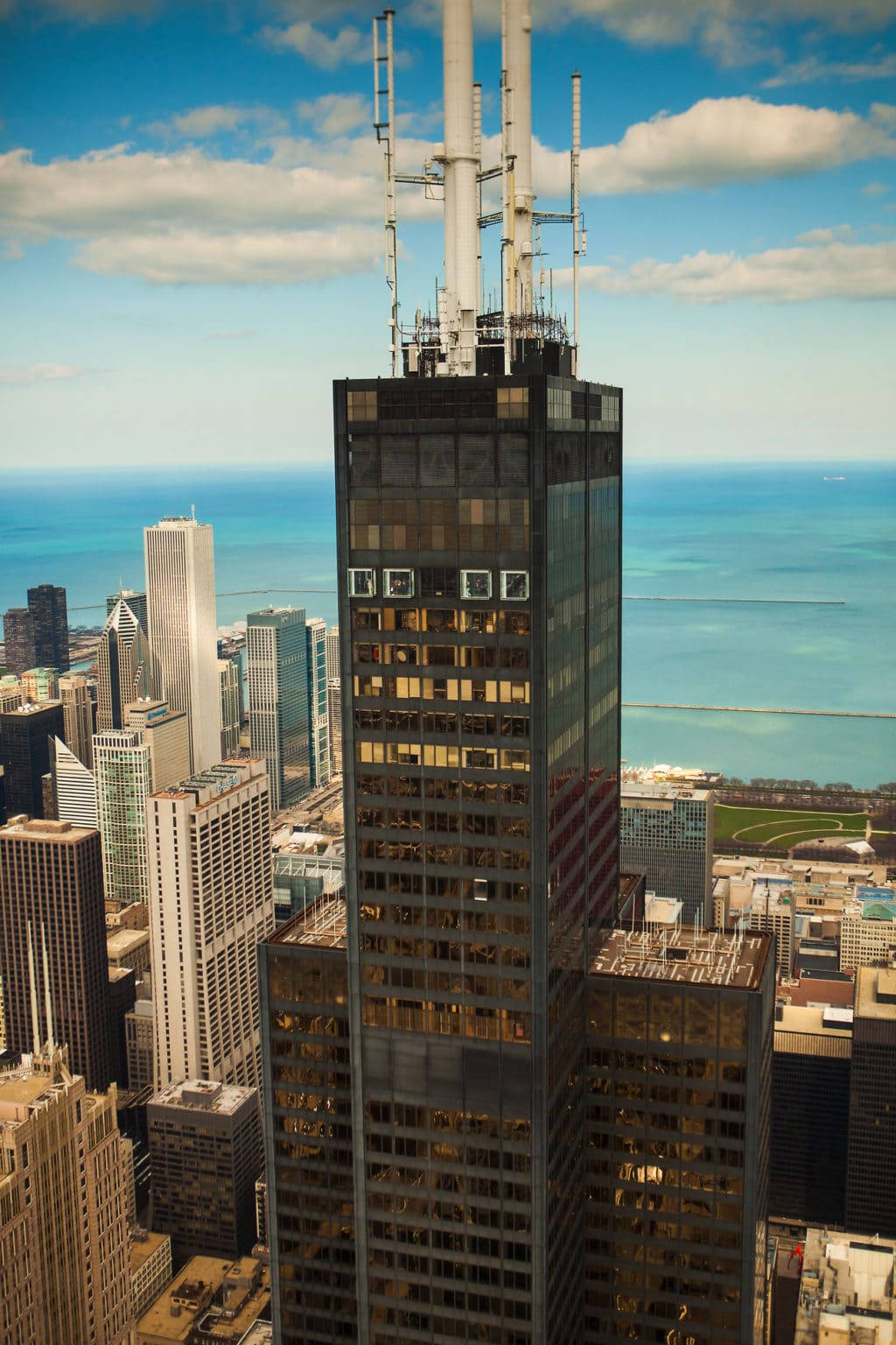 Buildings 桌布 壁紙 — 從高空俯瞰一座高聳入雲的摩天大樓，帶有天線陣列，俯瞰城市景觀和藍色湖泊，天空部分多雲。