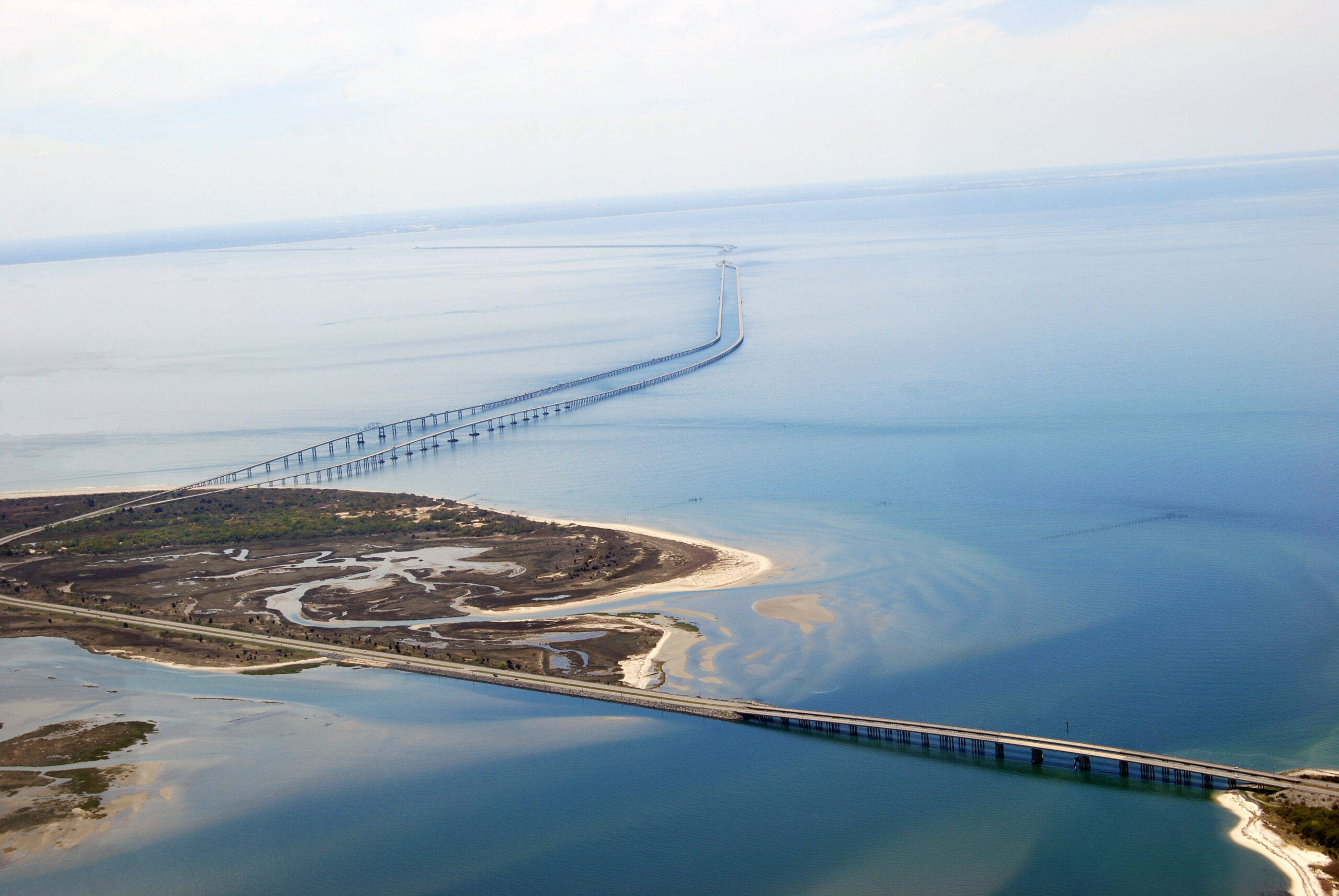 島嶼 桌布 壁紙 — 從高空俯瞰一條長橋，橫跨平靜的藍色水域，延伸至明亮天空下的遙遠地平線。