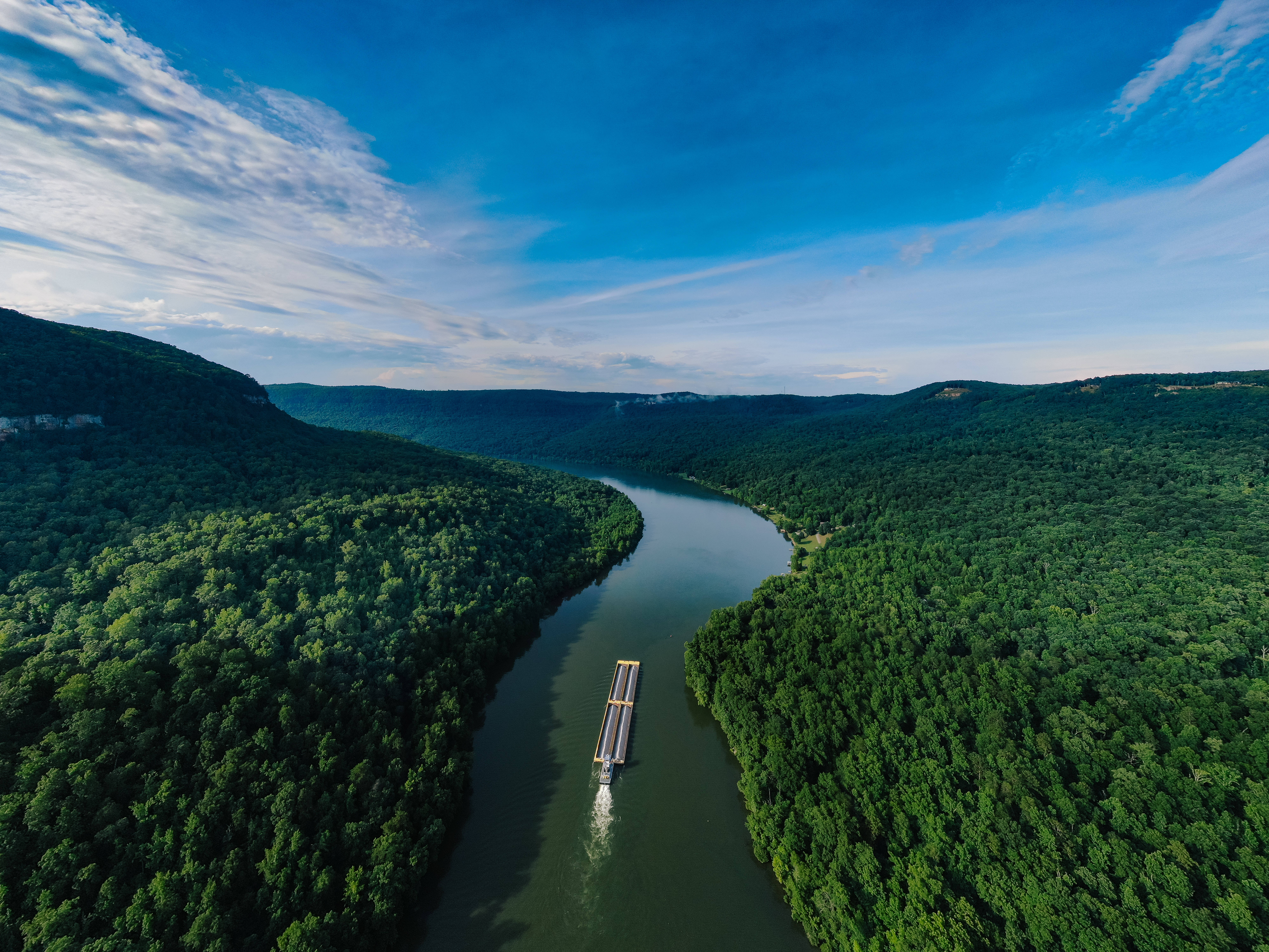 river 桌布 壁紙 — 從高空俯瞰一艘長長的駁船在蜿蜒的河流中航行，兩旁是茂密的綠色森林，上方是明亮的藍天，飄著縷縷白雲。