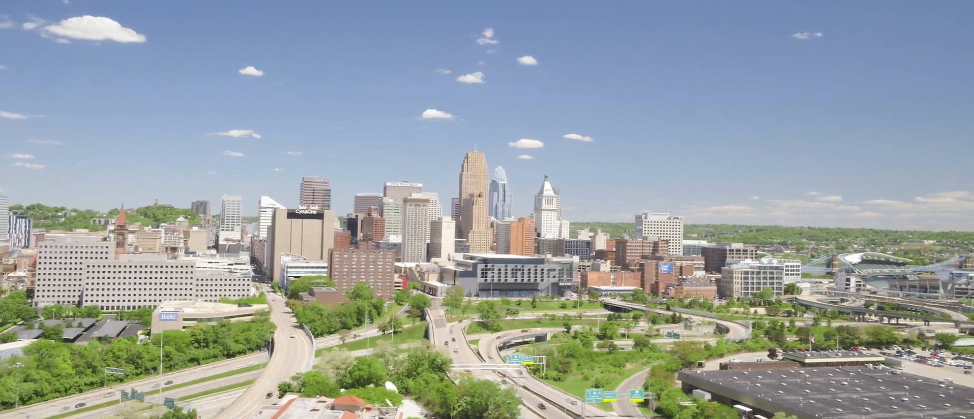 Cincinnati 桌布 壁紙 — 從高空俯瞰充滿活力的城市景觀，現代摩天大樓、綠色公園和蜿蜒的高速公路，在明亮的藍天和零星雲朵下。