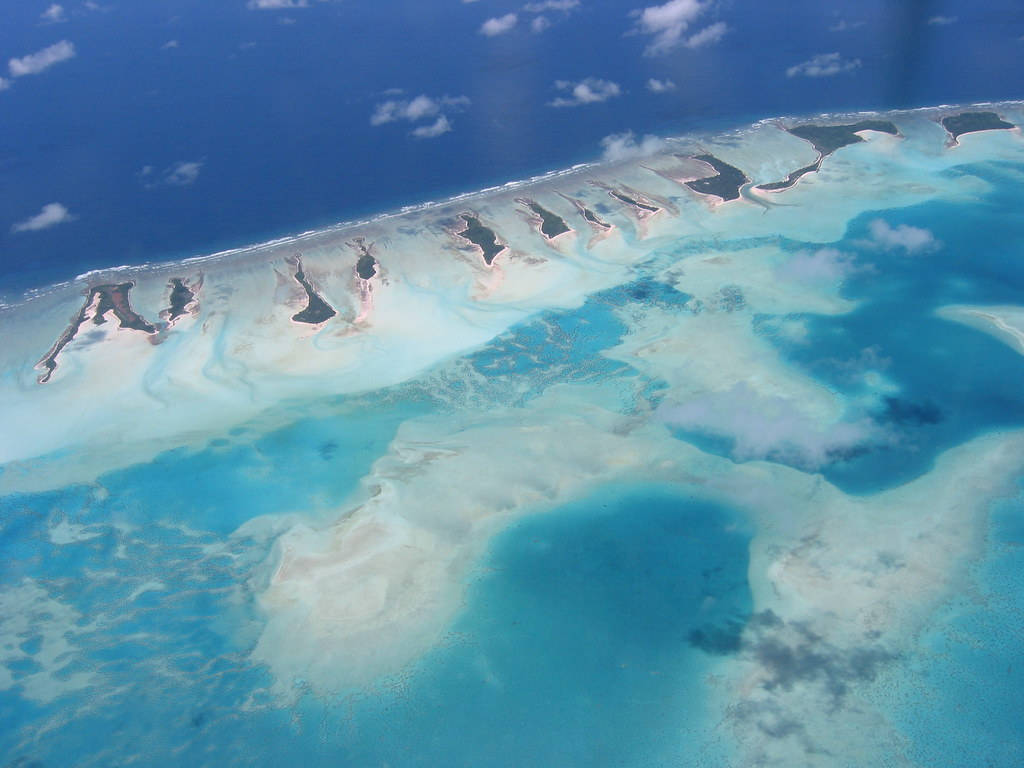 Kiribati 桌布 壁紙 — 從高空俯瞰壯麗環礁，碧綠潟湖與白色沙洲在深藍色海洋中蜿蜒，天空散落著雲朵。