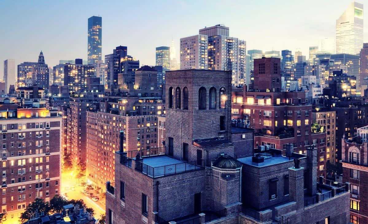 Buildings 桌布 壁紙 — 從高空俯瞰密集的城市天際線，在黃昏時分，溫暖的街燈和發光的窗戶映襯著柔和的藍天。