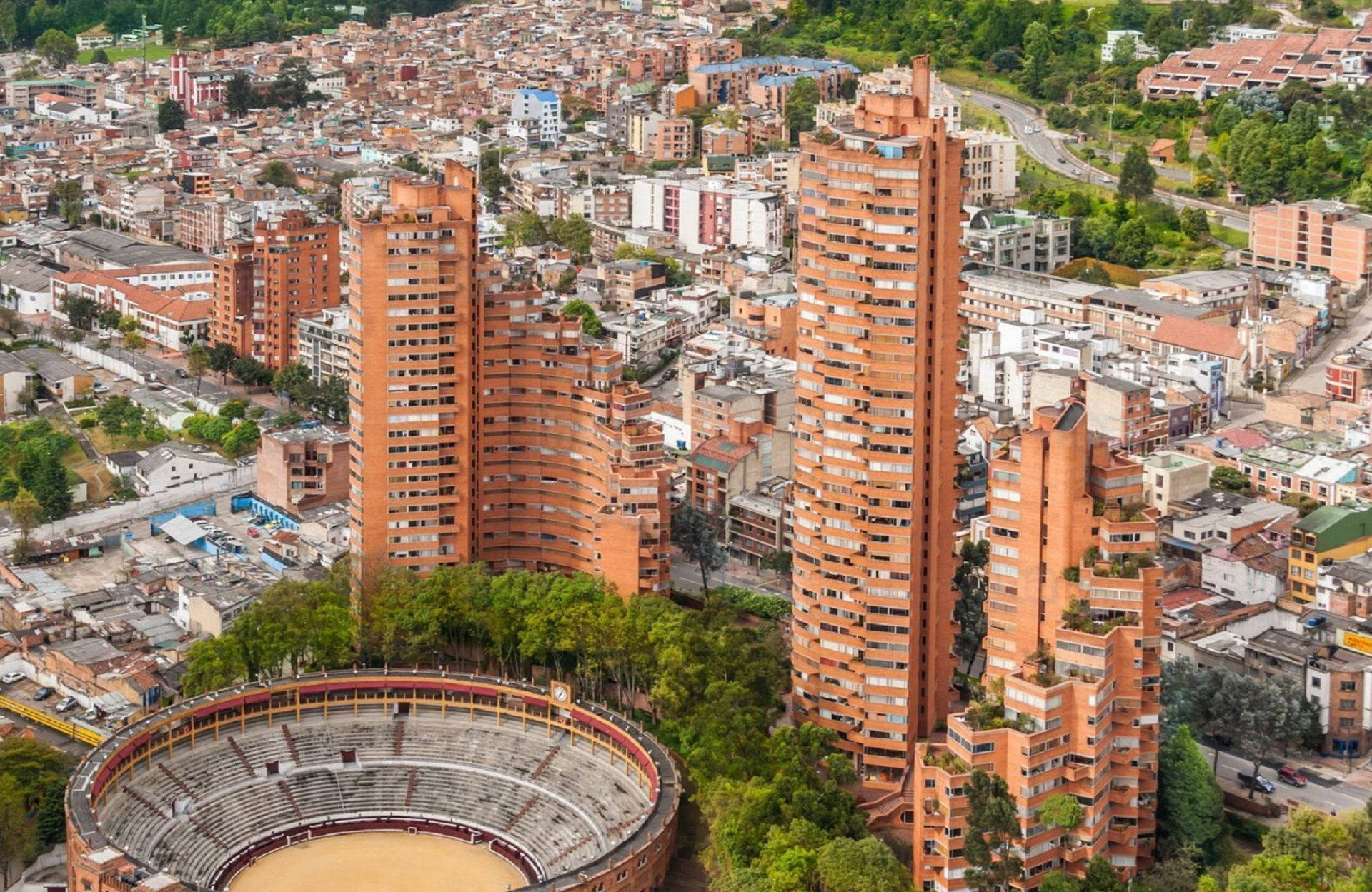 Bogota 桌布 壁紙 — 從高空俯瞰密集的城市景觀，以突出的陶土色高樓大廈和一個被綠樹環繞的圓形鬥牛場為特色。