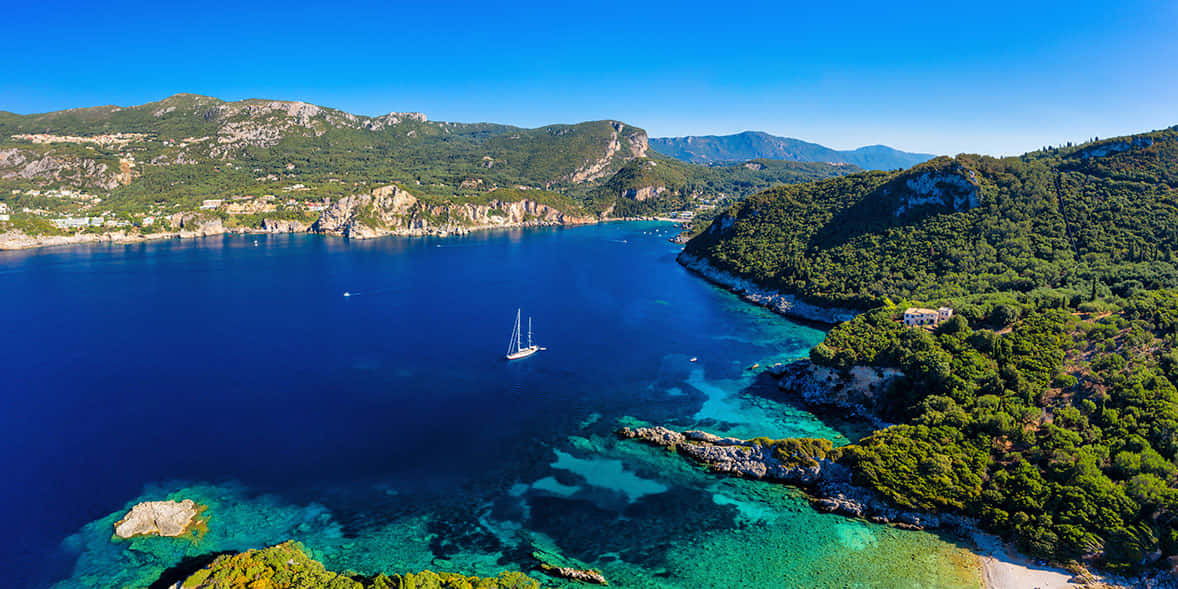 Greek Island 桌布 壁紙 — 從高空俯瞰寧靜的海岸海灣，碧綠的海水中，一艘孤獨的帆船在鬱鬱蔥蔥的綠色山丘之間滑行，天空湛藍清澈。