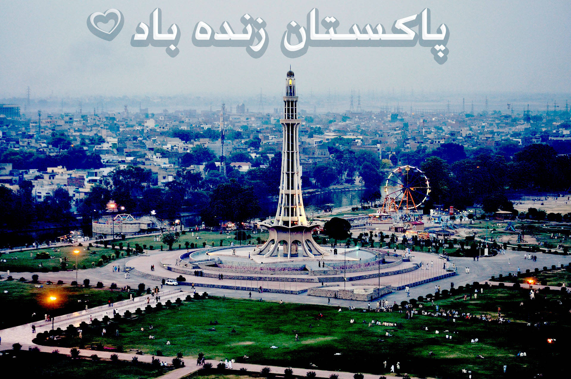 巴基斯坦 桌布 壁紙 — 從高空俯瞰廣闊公園中高聳的 Minar-e-Pakistan 紀念碑，摩天輪和城市景觀在朦朧的暮色中展現。