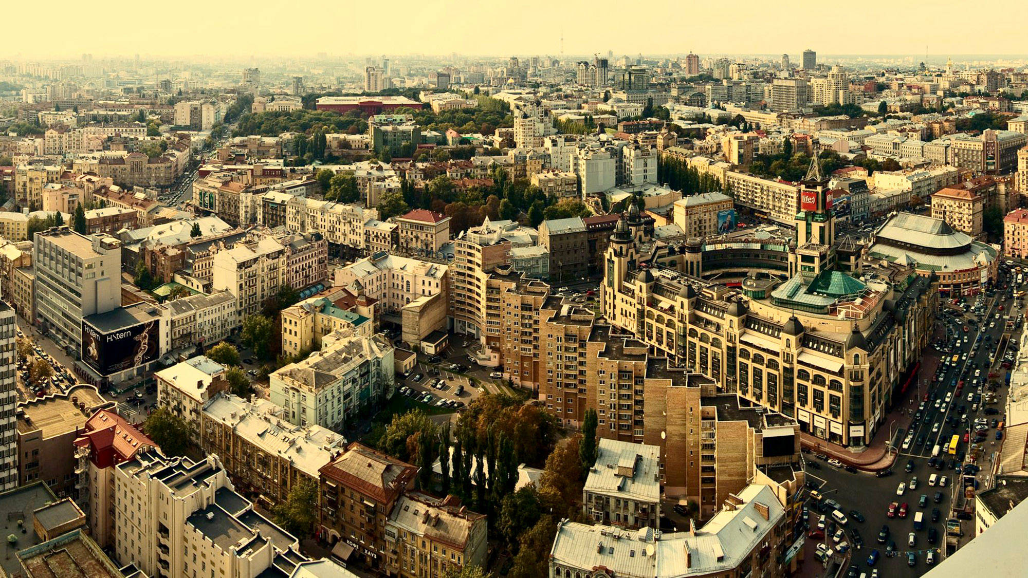 Ukraine 桌布 壁紙 — 從高空俯瞰廣闊的城市景觀，密集的建築物、蜿蜒的街道，以及一座在溫暖金色光線下顯著的華麗建築。