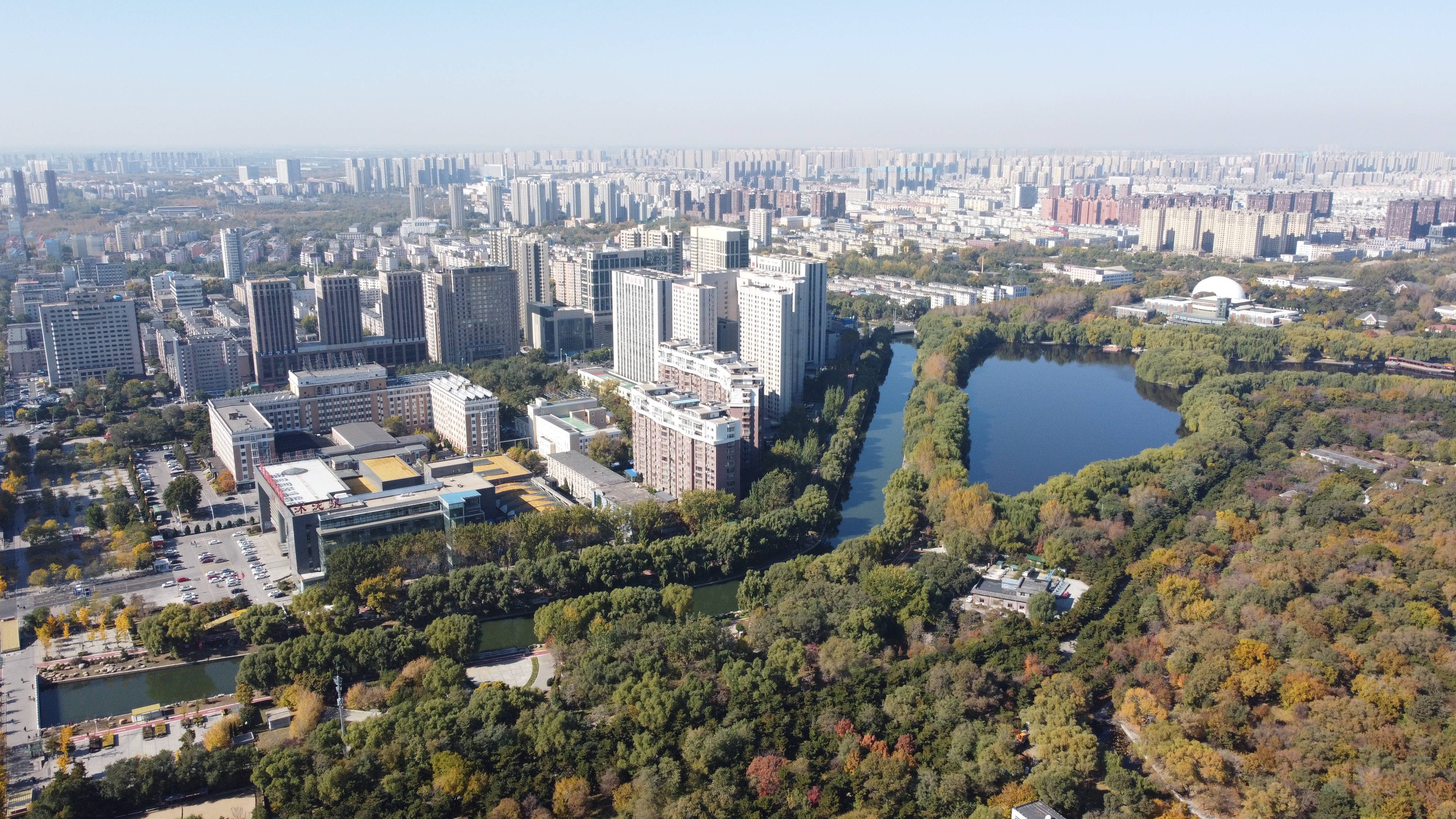 中國 桌布 壁紙 — 從高空俯瞰廣闊的城市景觀，現代高層建築坐落在秋色樹木之中，環繞著蜿蜒的河流和一個大湖。