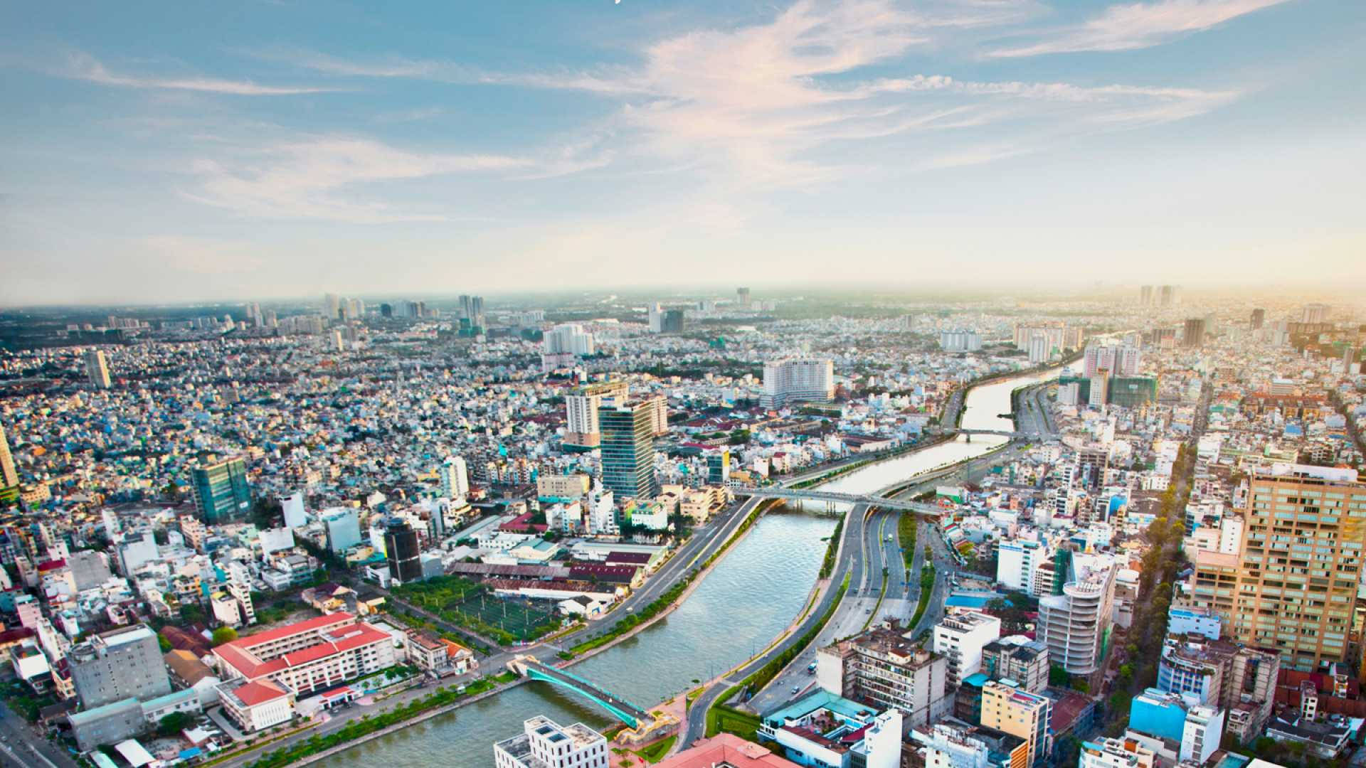Ho Chi Minh City 桌布 壁紙 — 從高空俯瞰廣闊的城市景觀，蜿蜒的河流穿過密集的城市建築，在柔和的藍天下。