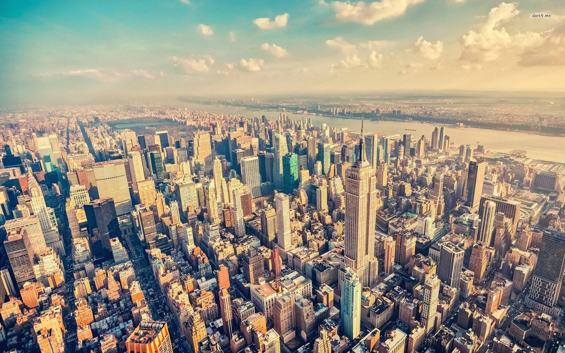 manhattan 桌布 壁紙 — 從高空俯瞰廣闊的城市景觀，高聳的摩天大樓在部分多雲的天空下沐浴在金色的陽光中。