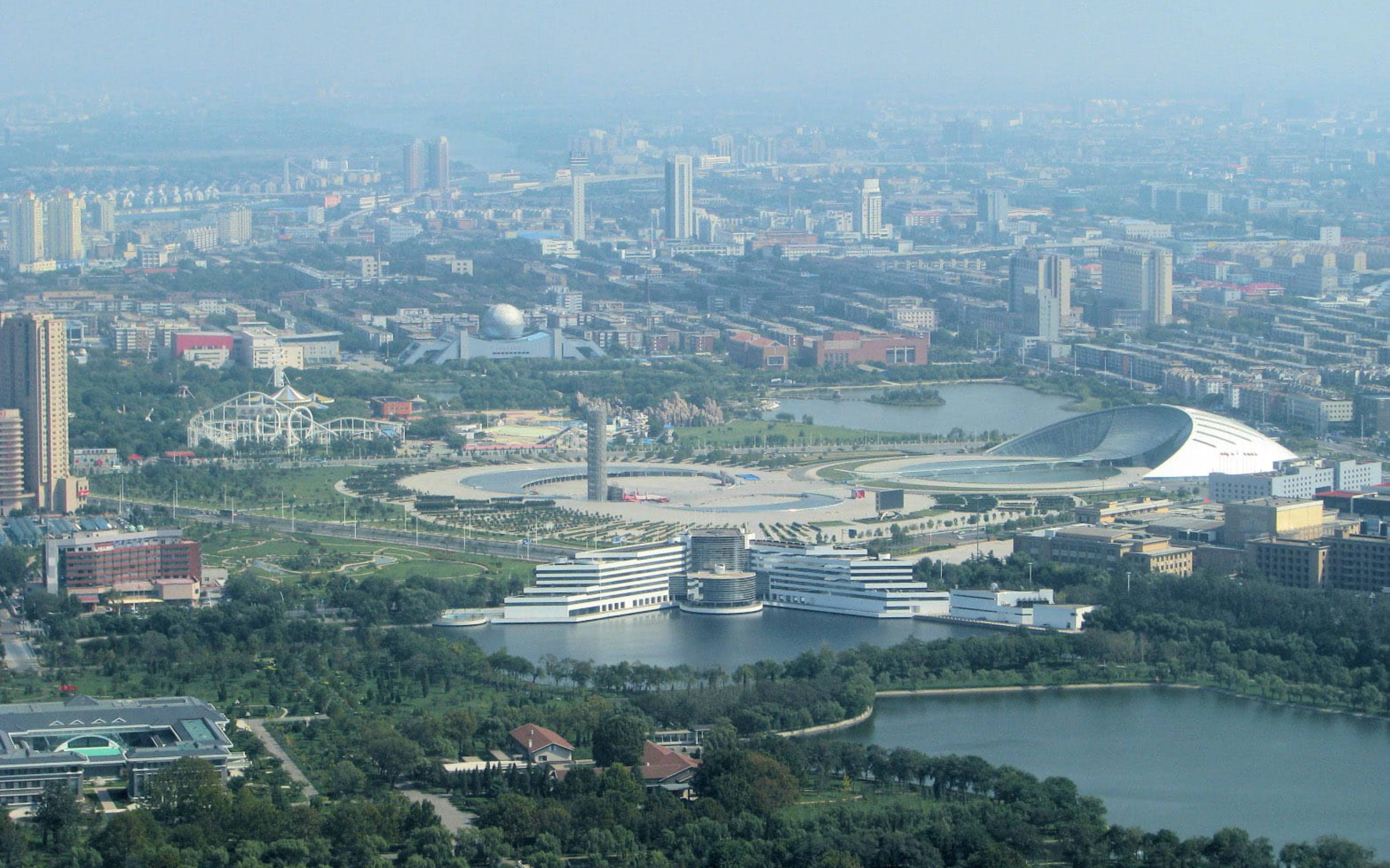 Tianjin 桌布 壁紙 — 從高空俯瞰廣闊的現代城市景觀，包含湖泊、綠色公園和未來主義建築，在朦朧的日光下。
