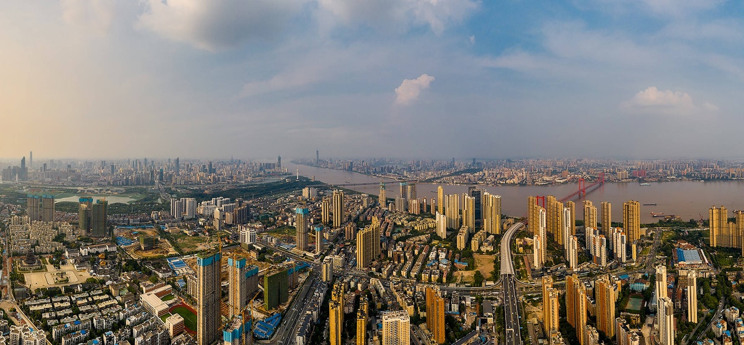 Wuhan 桌布 壁紙 — 從高空俯瞰廣闊的現代城市景觀，高樓大廈密集，道路蜿蜒，一條寬闊的河流上有一座紅色橋樑，天空部分多雲。
