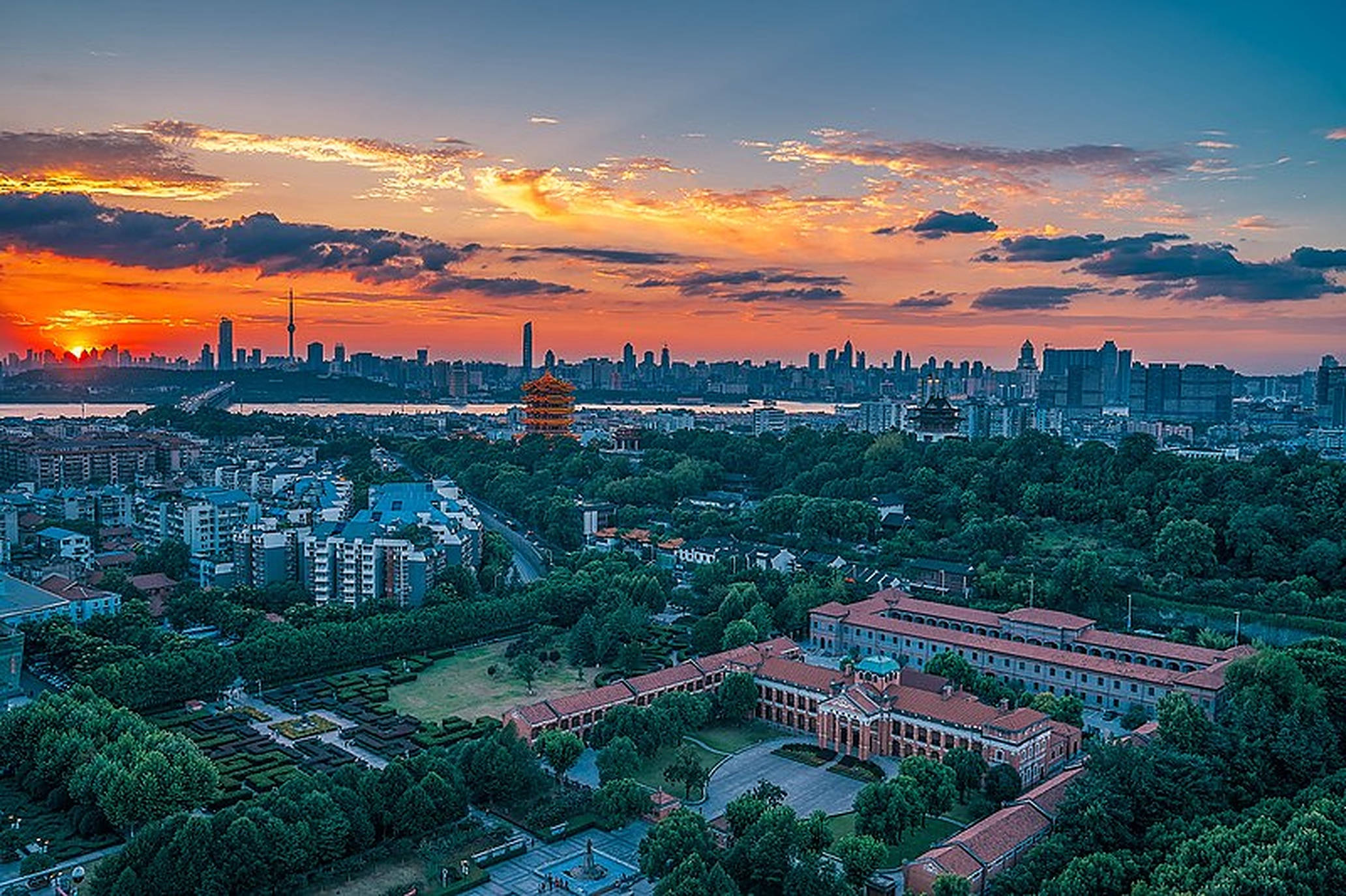 Wuhan 桌布 壁紙 — 從高空俯瞰日落時分的城市景觀，河流上方有橙色雲彩，歷史建築被茂密的綠樹環繞。