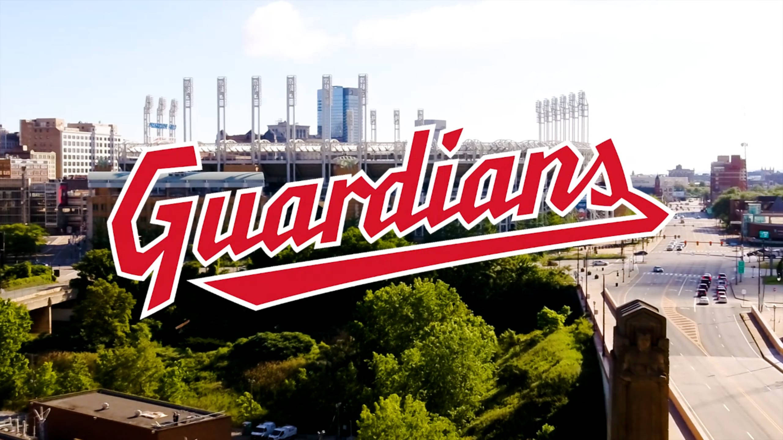 Cleveland Guardians 桌布 壁紙 — 從高空俯瞰棒球場，'Guardians' 標誌疊加在一個有綠樹和道路的城市景觀上，在明亮的日光下。