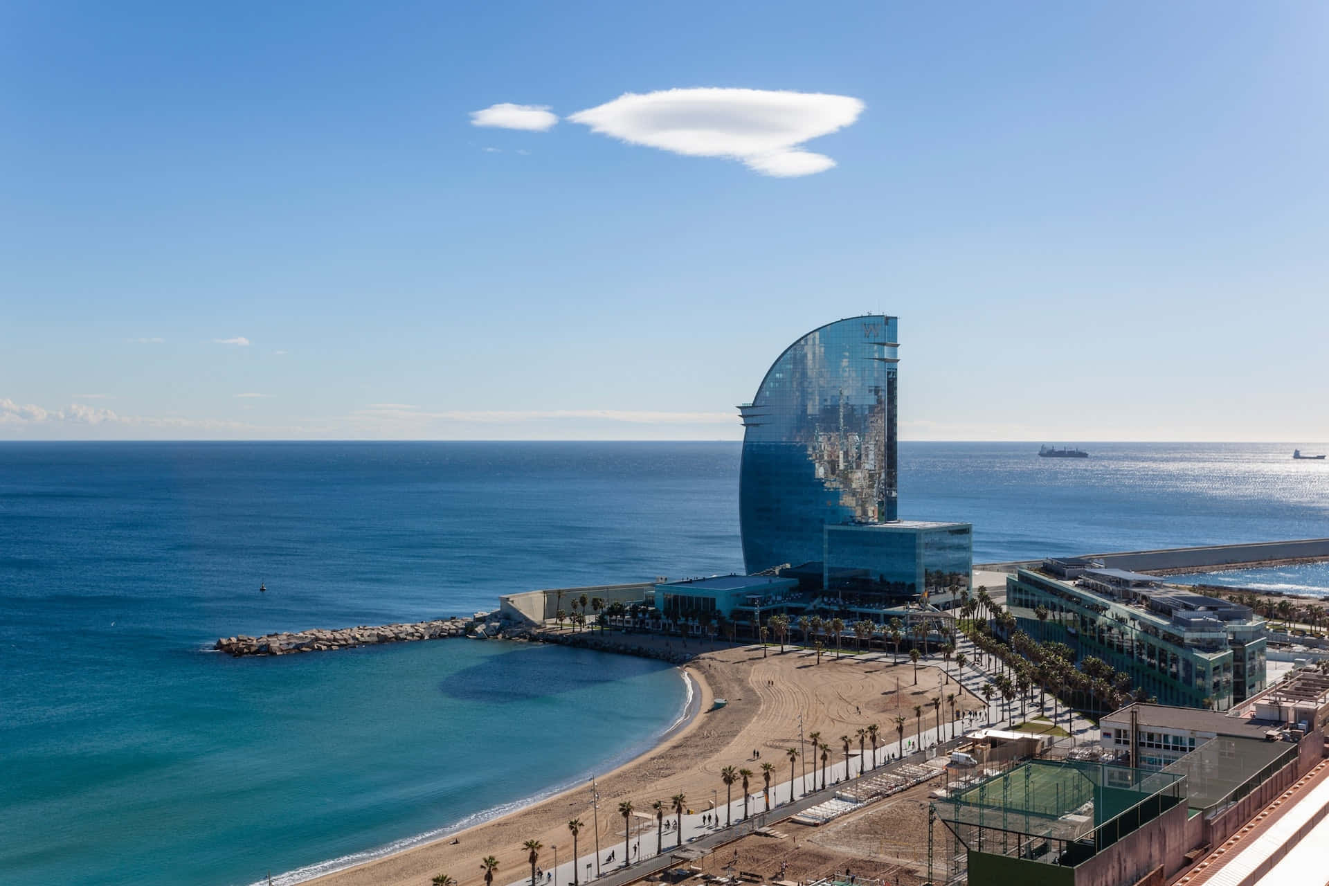 Barcelona 桌布 壁紙 — 從高空俯瞰標誌性W巴塞隆納酒店，在清澈藍天下，沙灘與碧綠海水旁，酒店玻璃幕牆倒映天空的壯麗景象。
