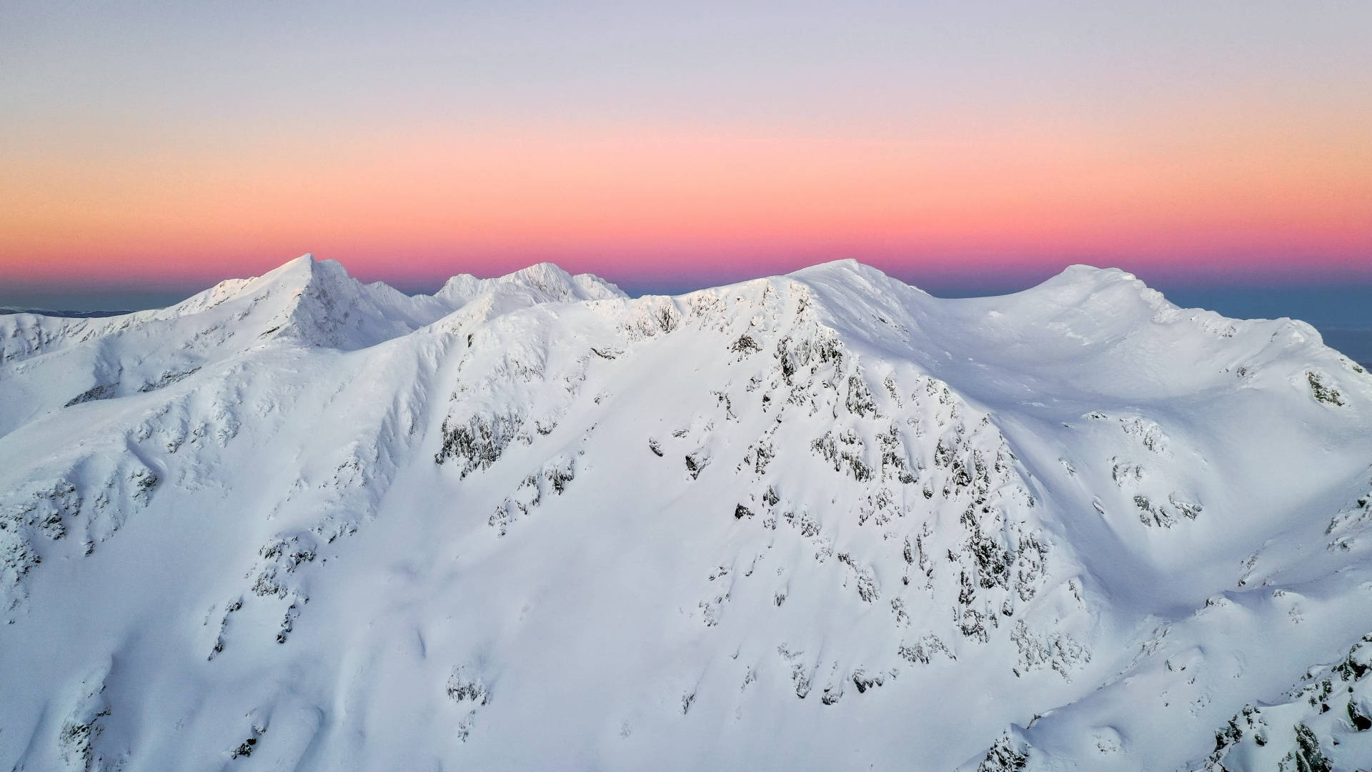 冬季美學 桌布 壁紙 — 從高空俯瞰白雪覆蓋的山峰，在晴朗的天空下沐浴在柔和的粉紅色和橙色日出色調中。