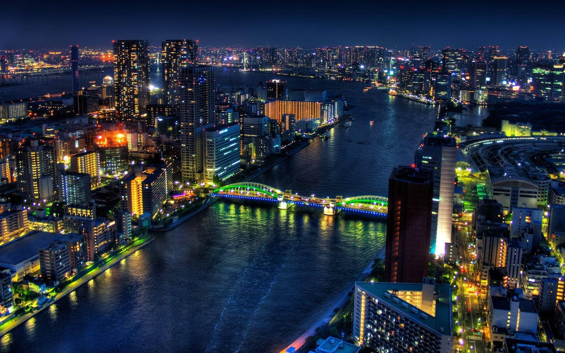東京 桌布 壁紙 — 從高空俯瞰的夜景，充滿活力的城市景觀，閃爍的摩天大樓和橫跨黑暗河流的彩色橋樑。