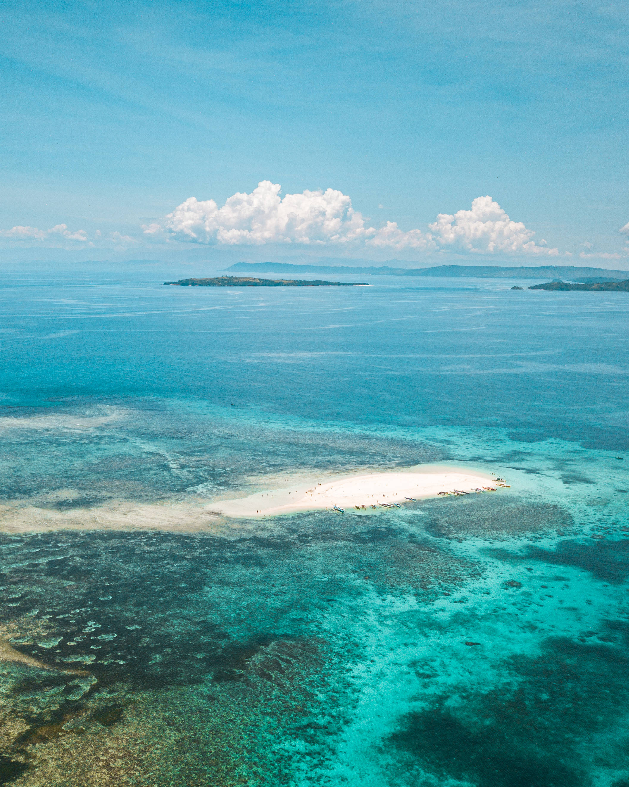 Siargao 桌布 壁紙 — 從高空俯瞰純白沙洲，周圍環繞著充滿活力的碧綠海水和珊瑚礁，上方是明亮的藍天和蓬鬆的白雲。