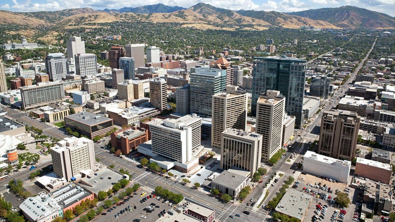 Salt Lake City 桌布 壁紙 — 從高空俯瞰繁華的城市景觀，現代摩天大樓和低層建築坐落在綠色公園和遠處乾旱山脈之間，天空湛藍明亮。