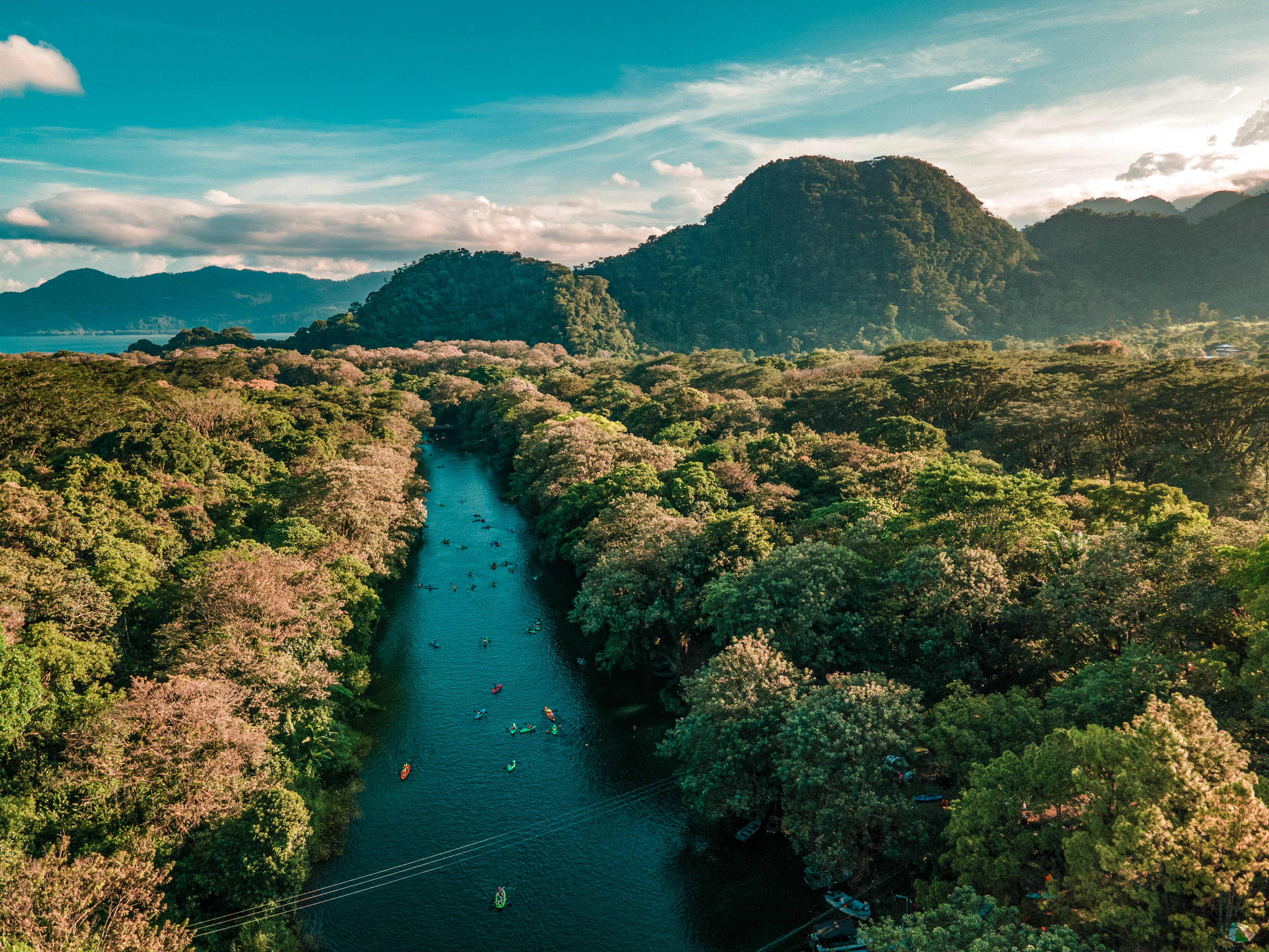 Honduras 桌布 壁紙 — 從高空俯瞰蜿蜒河流穿過茂密綠色森林，有獨木舟划手和遠處山脈，在明亮的藍天下。