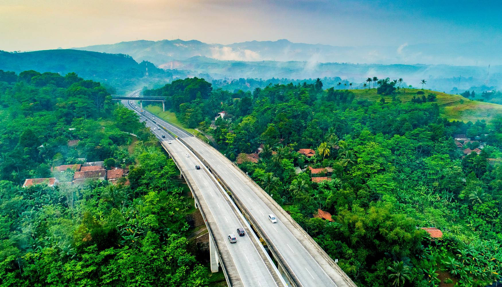 bandung 桌布 壁紙 — 從高空俯瞰蜿蜒的高速公路穿過茂密的綠色叢林，遠處有霧氣繚繞的山巒。