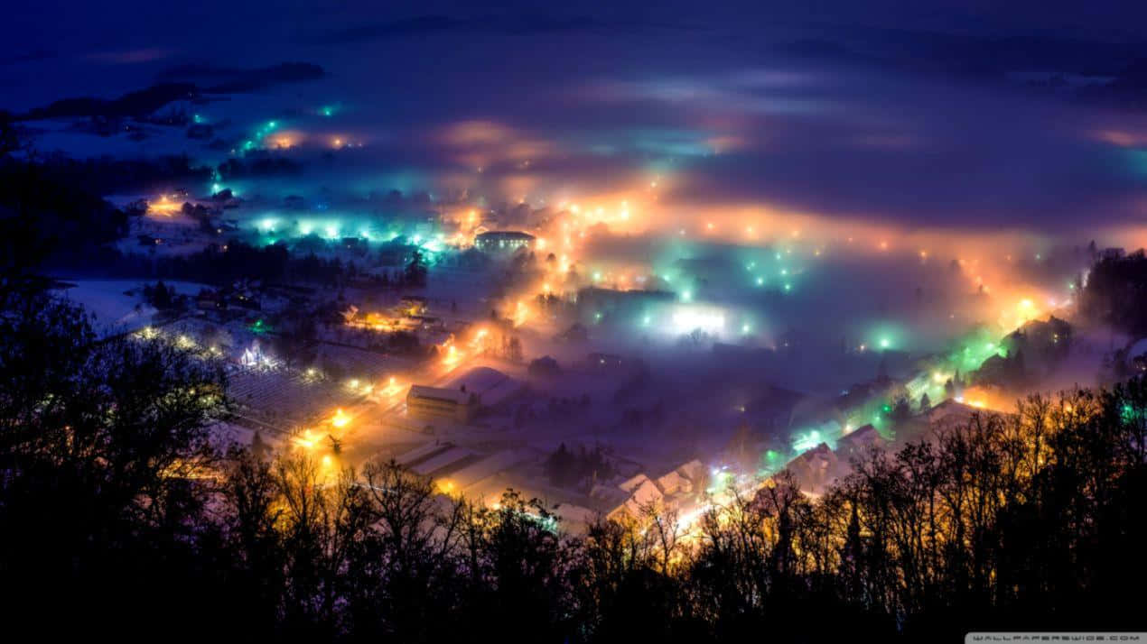 Winter Night Desktop 桌布 壁紙 — 從高處俯瞰，一個被雪覆蓋的村莊在夜晚沐浴在充滿活力的橙色和藍綠色燈光中，穿過霧氣。