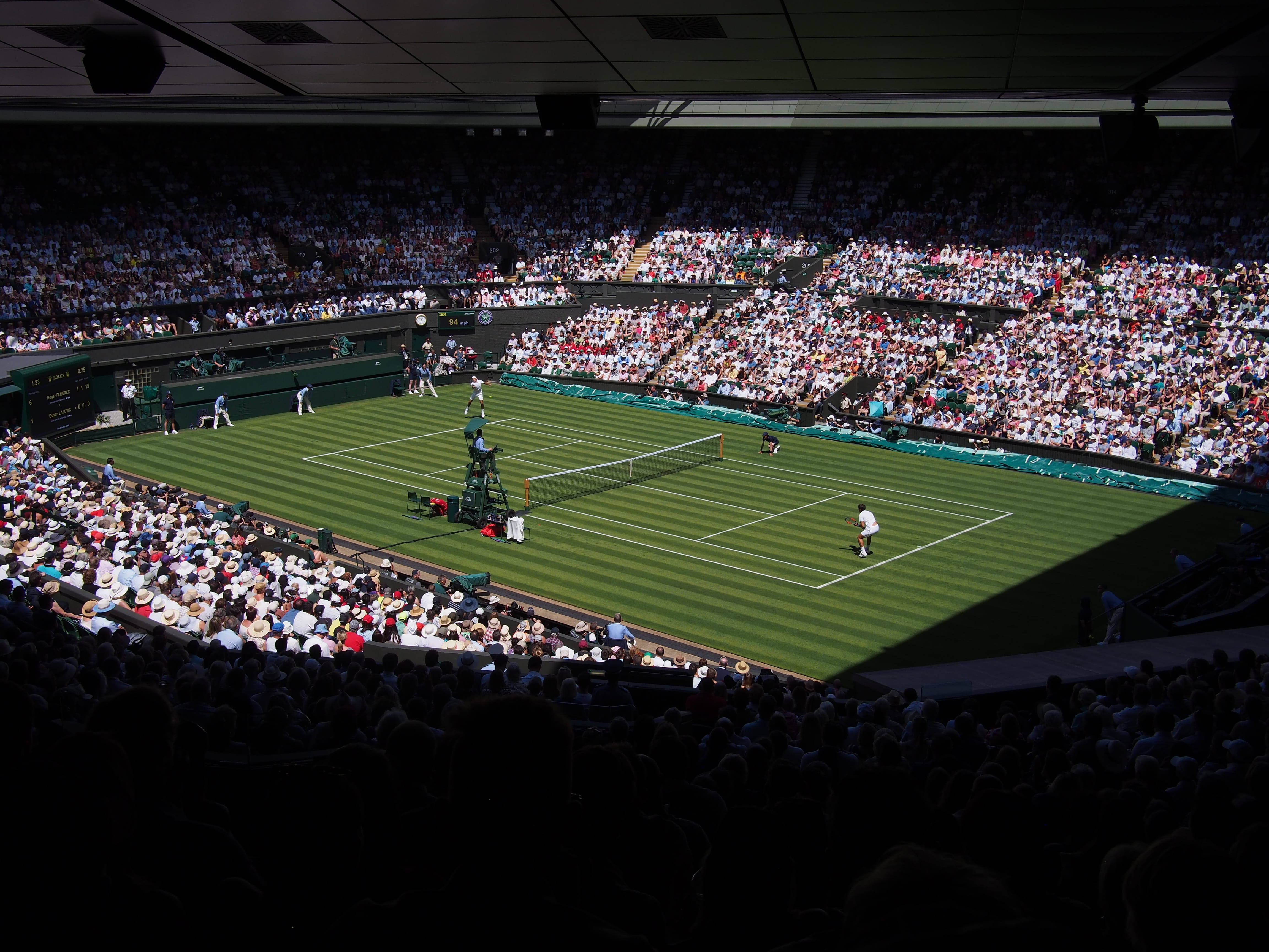 Wimbledon 桌布 壁紙 — 從高角度俯瞰溫布頓陽光普照的草地網球場，場上有球員和坐滿觀眾的看台。