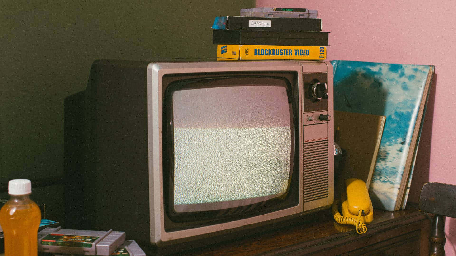 tv 桌布 壁紙 — 復古CRT電視機顯示靜態畫面，放在木架上，周圍有VHS錄影帶、遊戲卡帶和一部黃色轉盤電話。