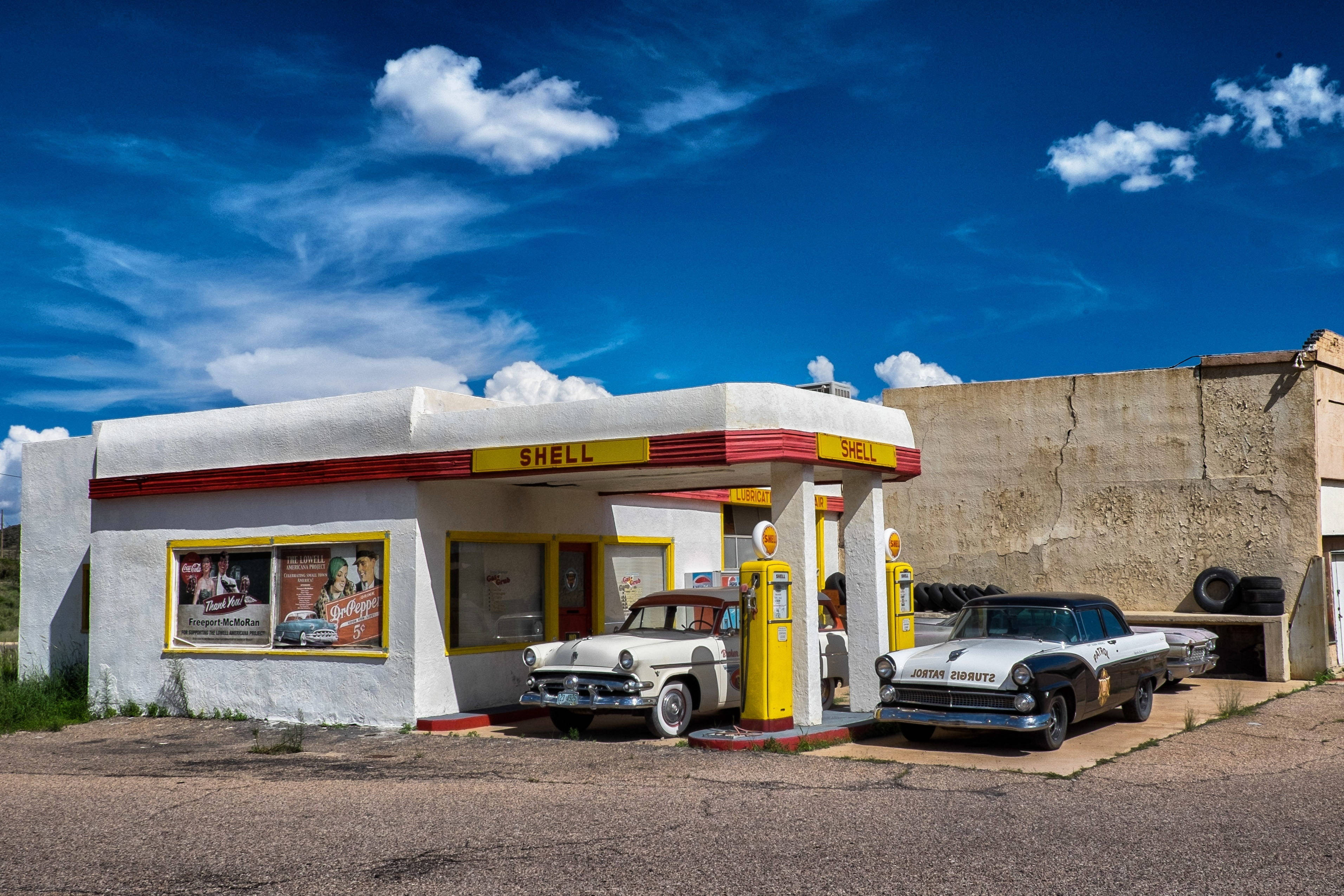 Gas Station 桌布 壁紙 — 復古Shell加油站，藍天白雲下，兩輛經典汽車停在黃色油泵旁。