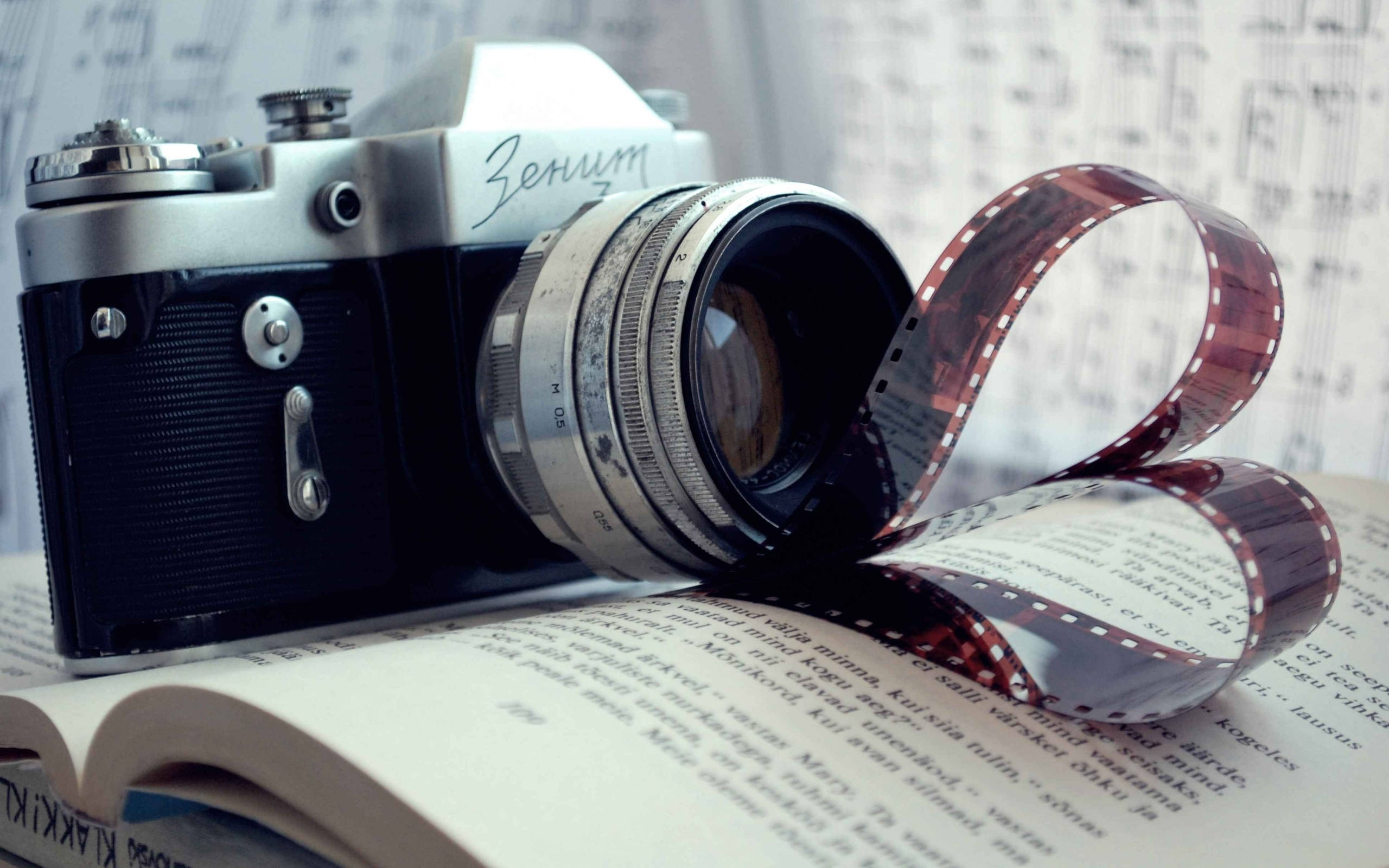 Dslr Camera 桌布 壁紙 — 復古Zenit相機擱在打開的書本上，底片卷曲如絲帶，襯以模糊的樂譜背景。